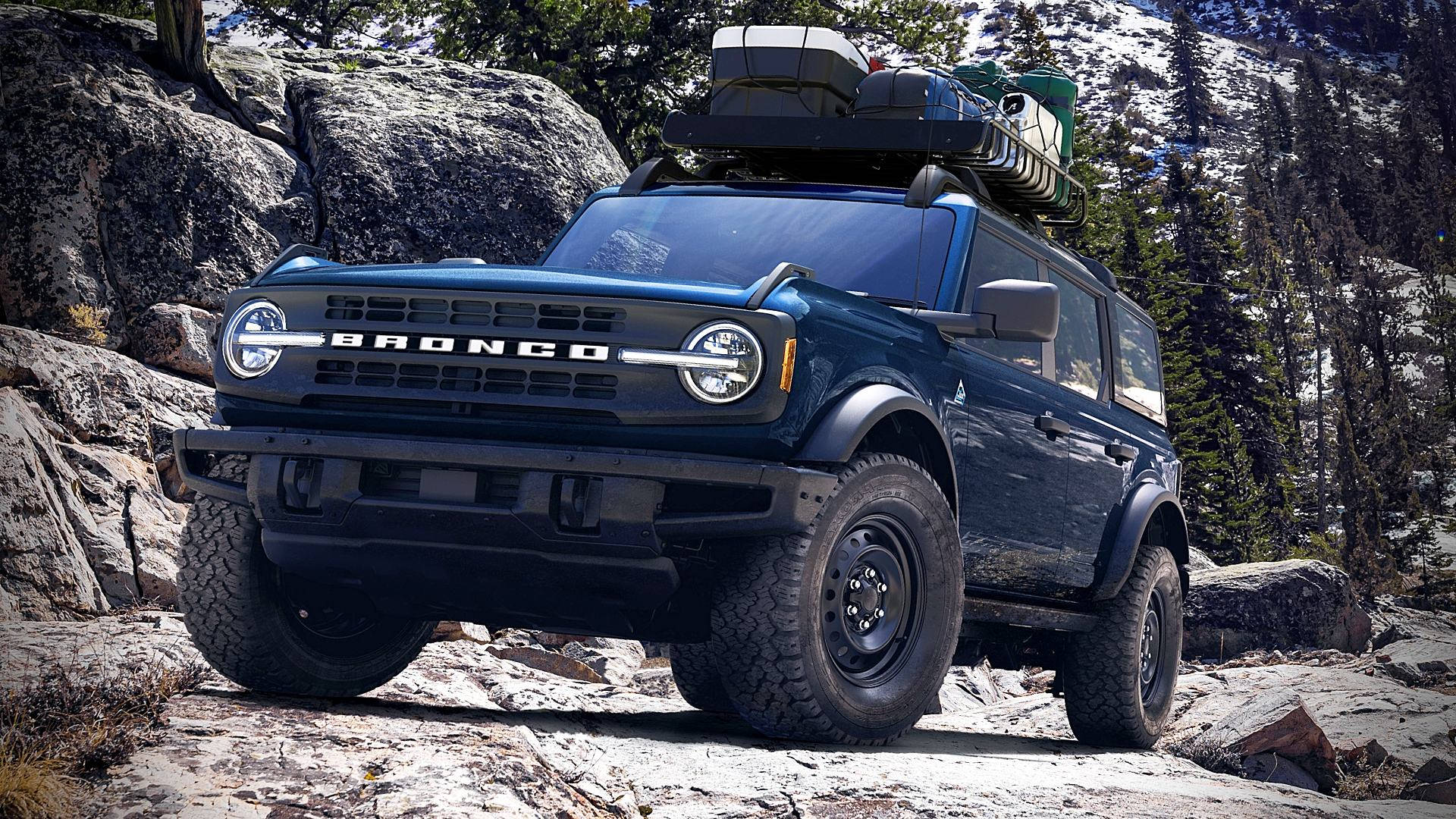 Ford Bronco 桌布 壁紙 — 一台堅固的藍色福特Bronco車頂載著貨物，停在白雪覆蓋的山岩上，周圍是常青樹。