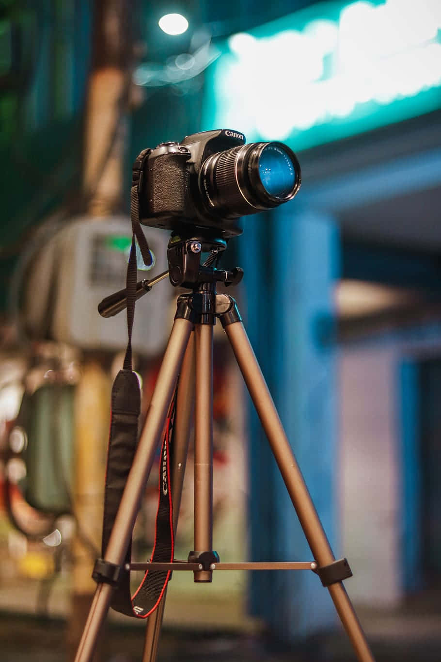 Dslr Camera 桌布 壁紙 — 一台安裝在三腳架上的黑色 Canon DSLR 相機，在充滿活力的霓虹燈城市夜景背景下蓄勢待發。