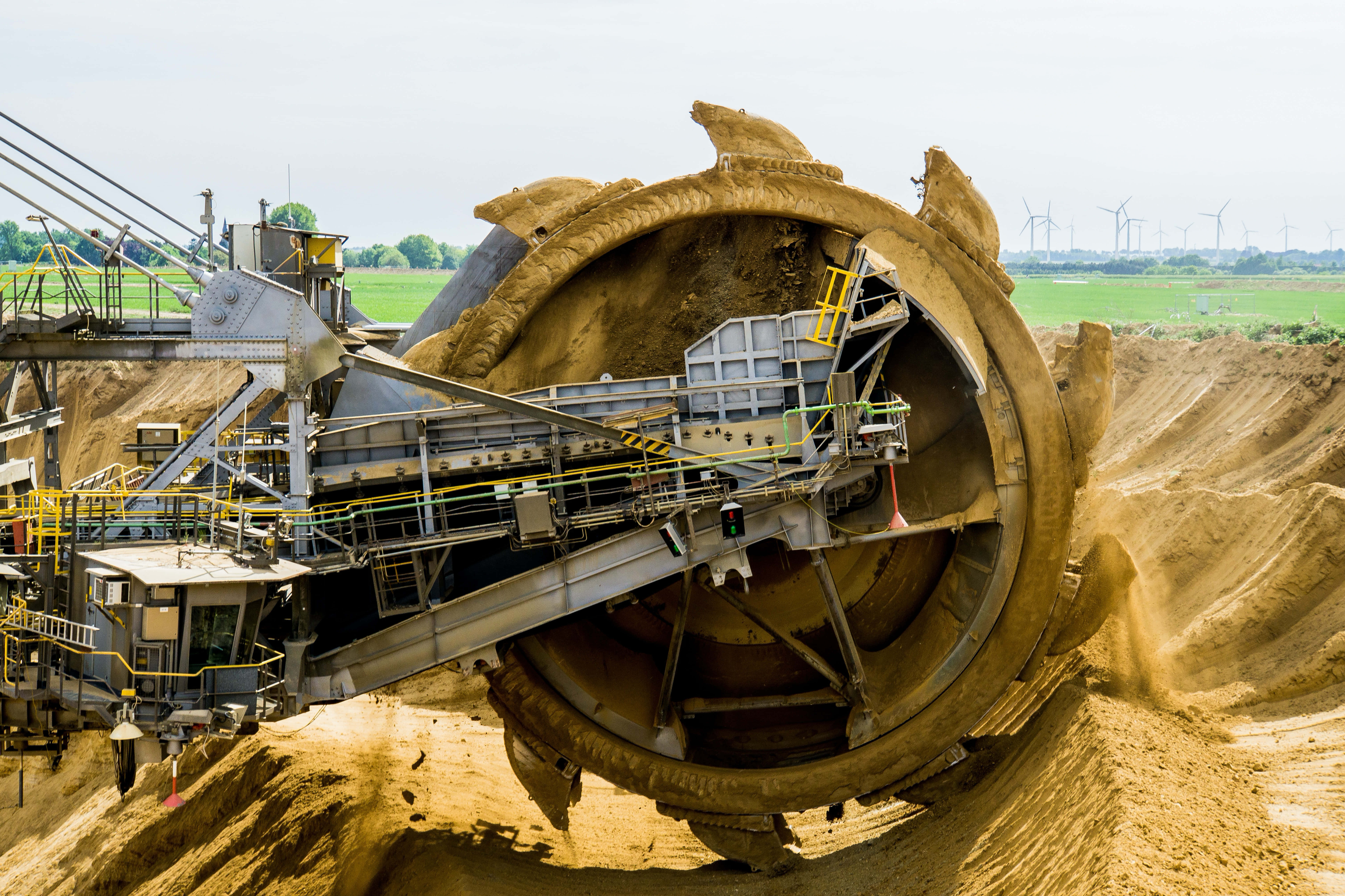 Mining 桌布 壁紙 — 一台巨大的斗輪挖掘機主宰著廣闊的露天礦場，天空明亮，遠處可見風力發電機。