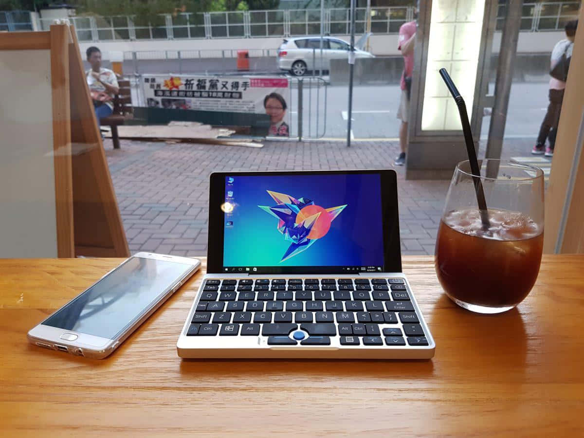 筆電 桌布 壁紙 — 一台帶有彩色摺紙鳥壁紙的緊湊型筆記型電腦放在咖啡廳的木桌上，旁邊是一部智慧型手機和冰咖啡。