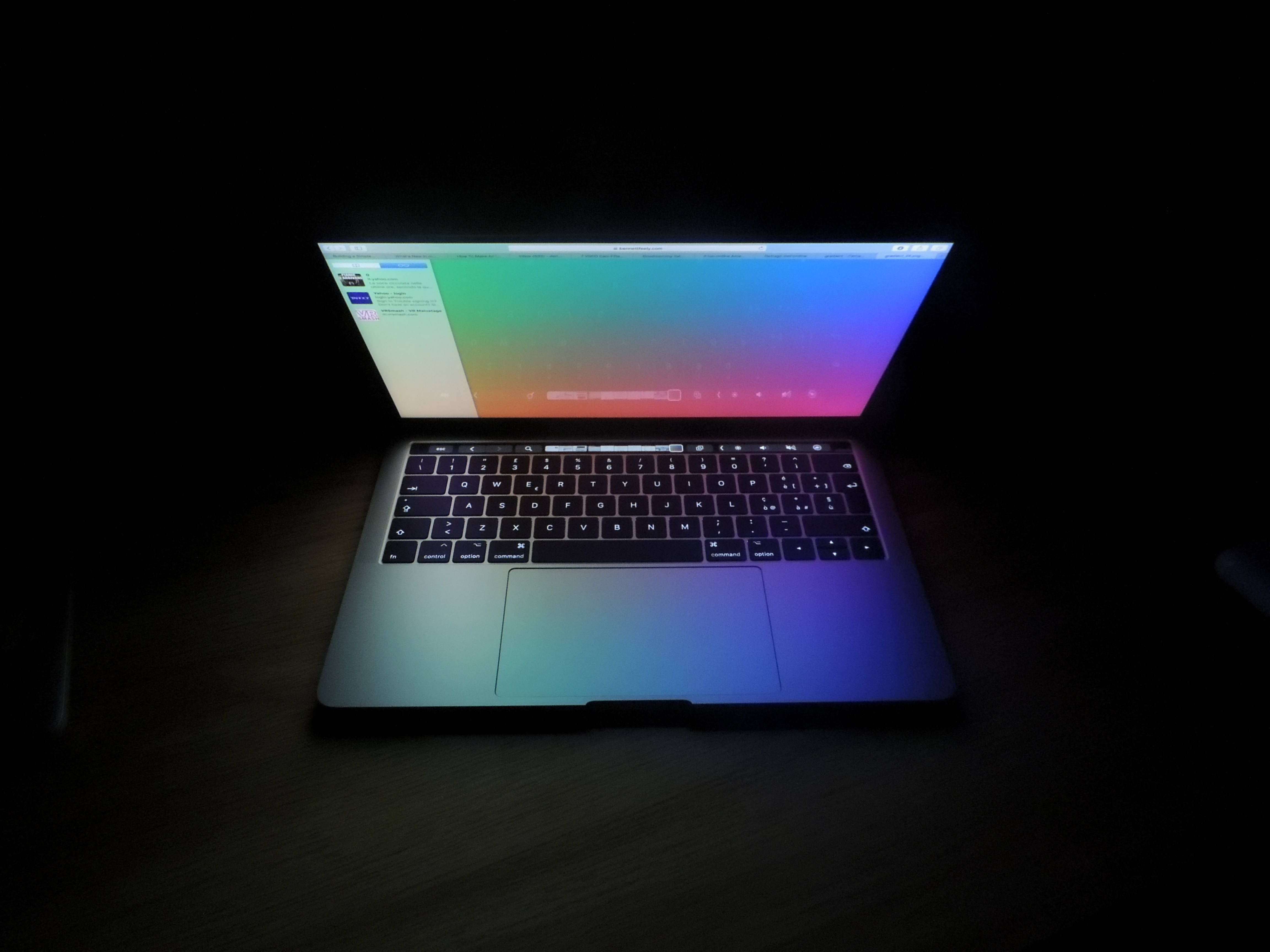 Dark Screen 桌布 壁紙 — 一台帶有鮮豔漸層螢幕的筆記型電腦在昏暗的房間中打開，放在深色木桌上。