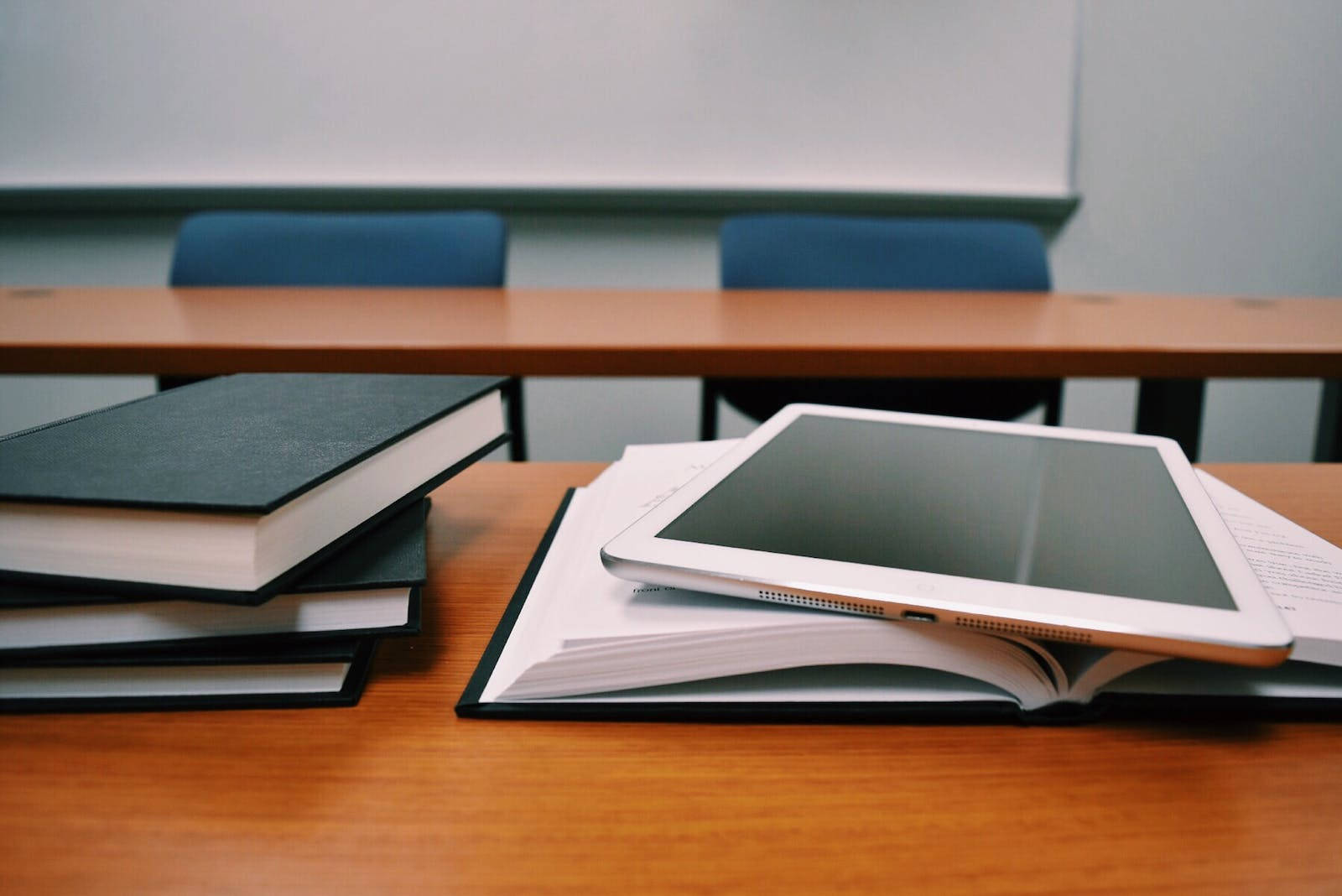 Education 桌布 壁紙 — 一台平板電腦放在木桌上的一本打開的書上，教室裡有椅子及白板，背景柔和模糊。
