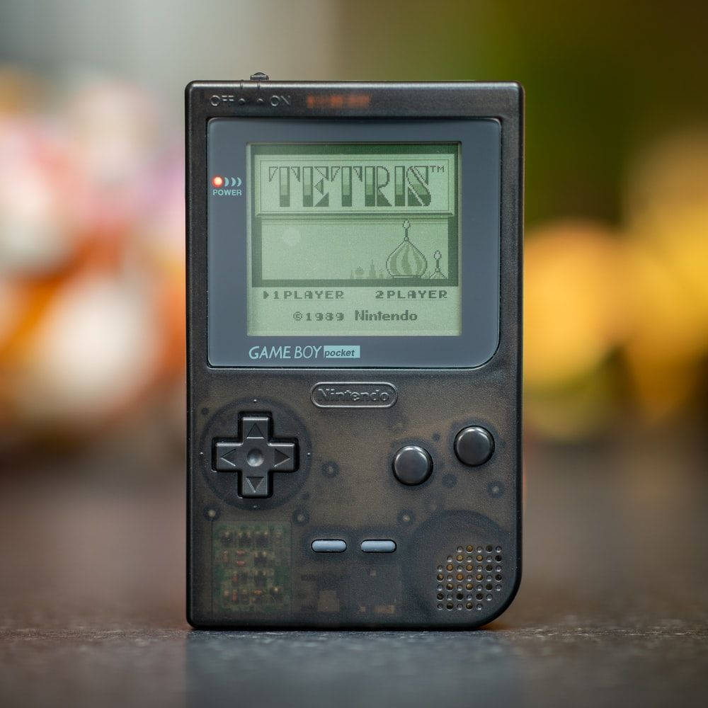 game 桌布 壁紙 — 一台復古 Game Boy Pocket 在模糊彩色背景上顯示經典俄羅斯方塊遊戲。