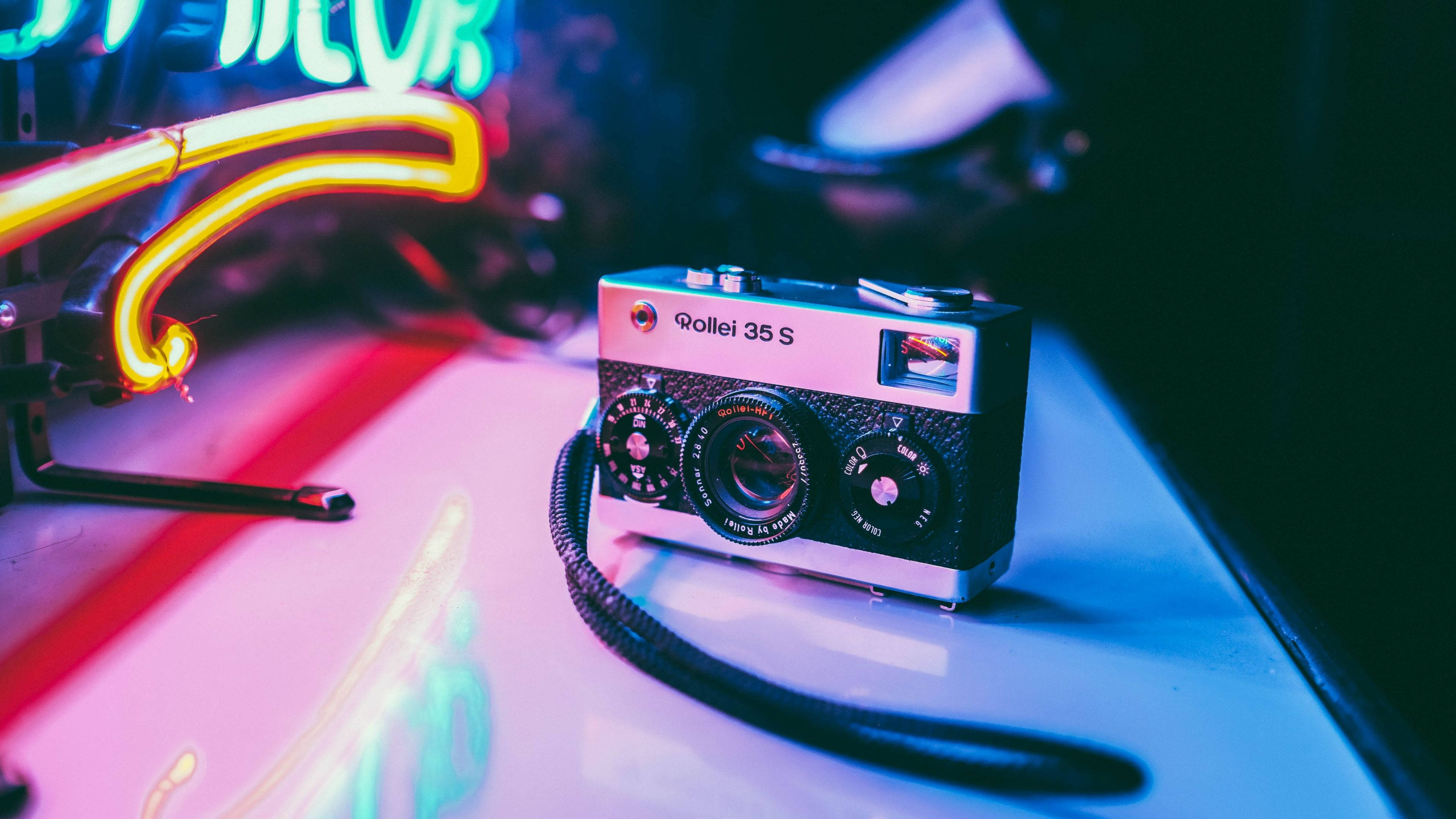 Retro Aesthetic 桌布 壁紙 — 一台復古 Rollei 35 S 相機放置在光滑表面上，沐浴在充滿活力的霓虹燈光中，投射出色彩繽紛的倒影。