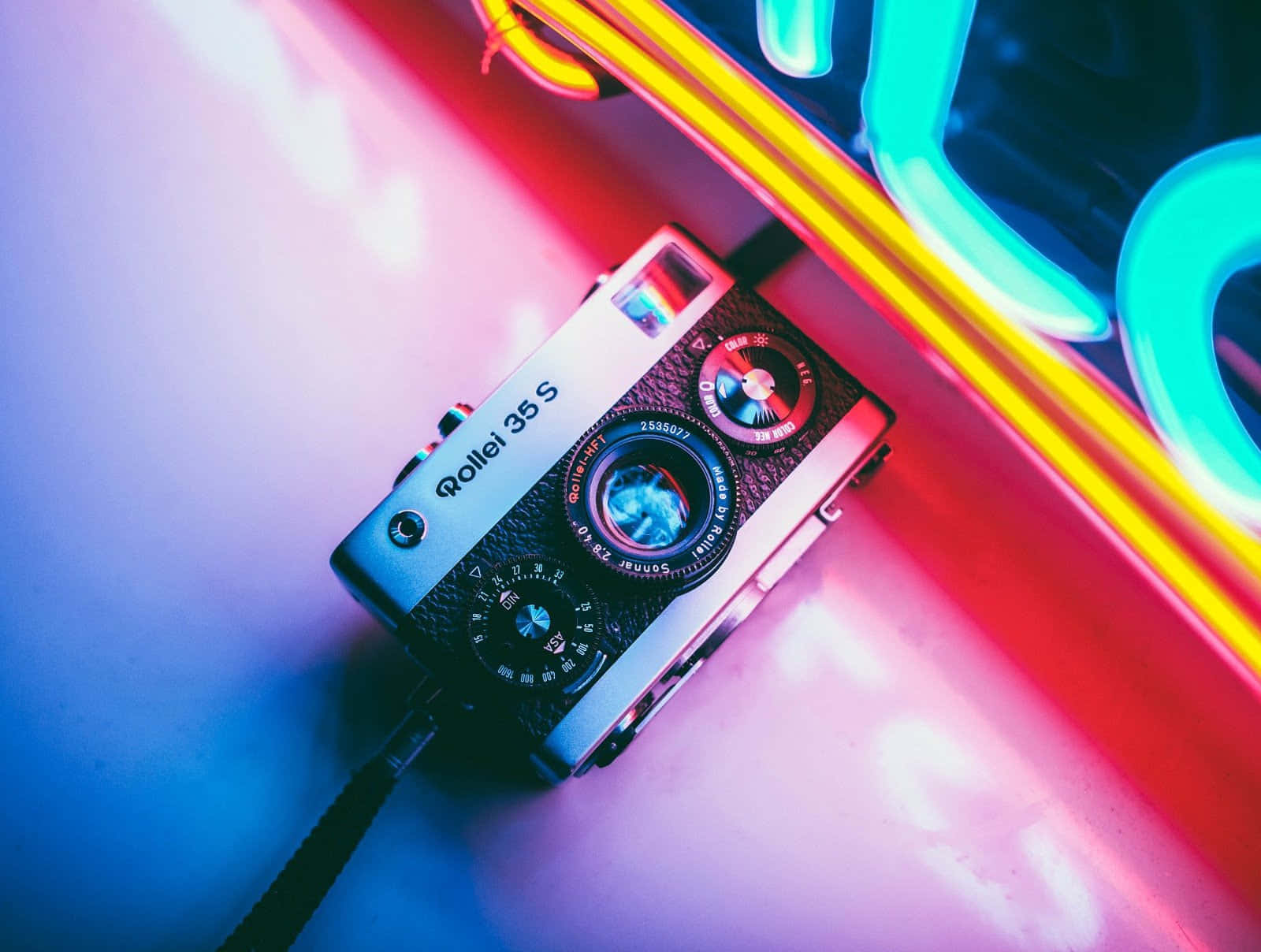 相機 桌布 壁紙 — 一台復古 Rollei 35S 相機放置在充滿活力的粉紅色和青色霓虹燈下，表面反射出色彩繽紛的光影。