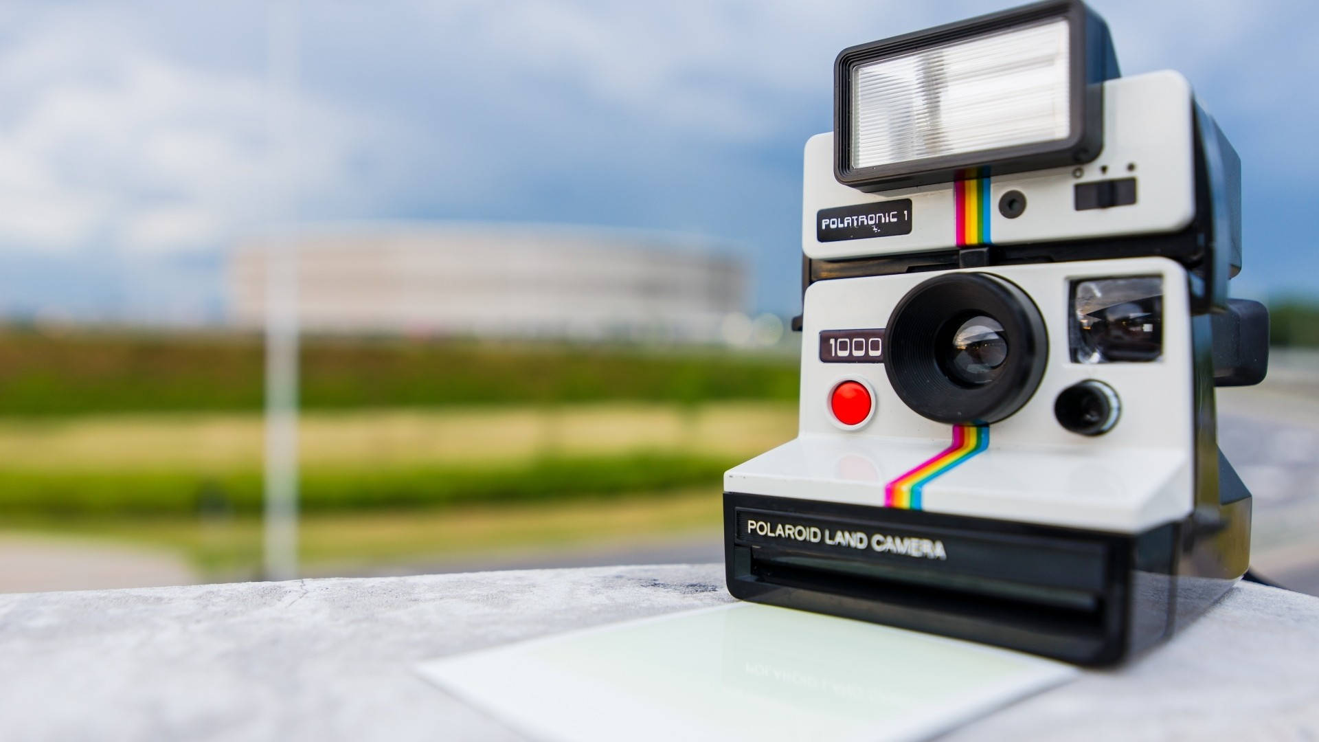 拍立得 桌布 壁紙 — 一台復古Polaroid Land Camera 1000相機放置在石頭表面上，背景是模糊的戶外景色，天空多雲。