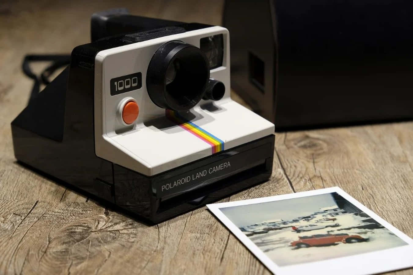 拍立得 桌布 壁紙 — 一台復古Polaroid Land Camera 1000相機放在木頭上，旁邊有一張雪中汽車的照片。
