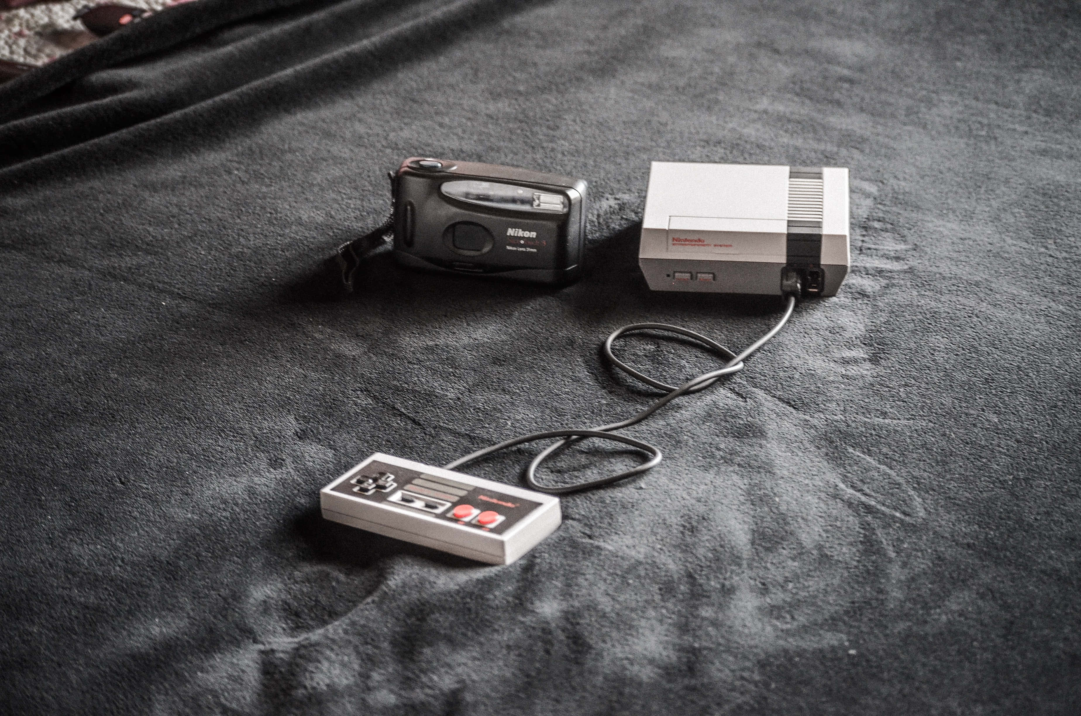 Gaming Consoles 桌布 壁紙 — 一台復古任天堂娛樂系統主機和控制器，與一台輕巧數位相機一同放在深色紋理布料表面上。