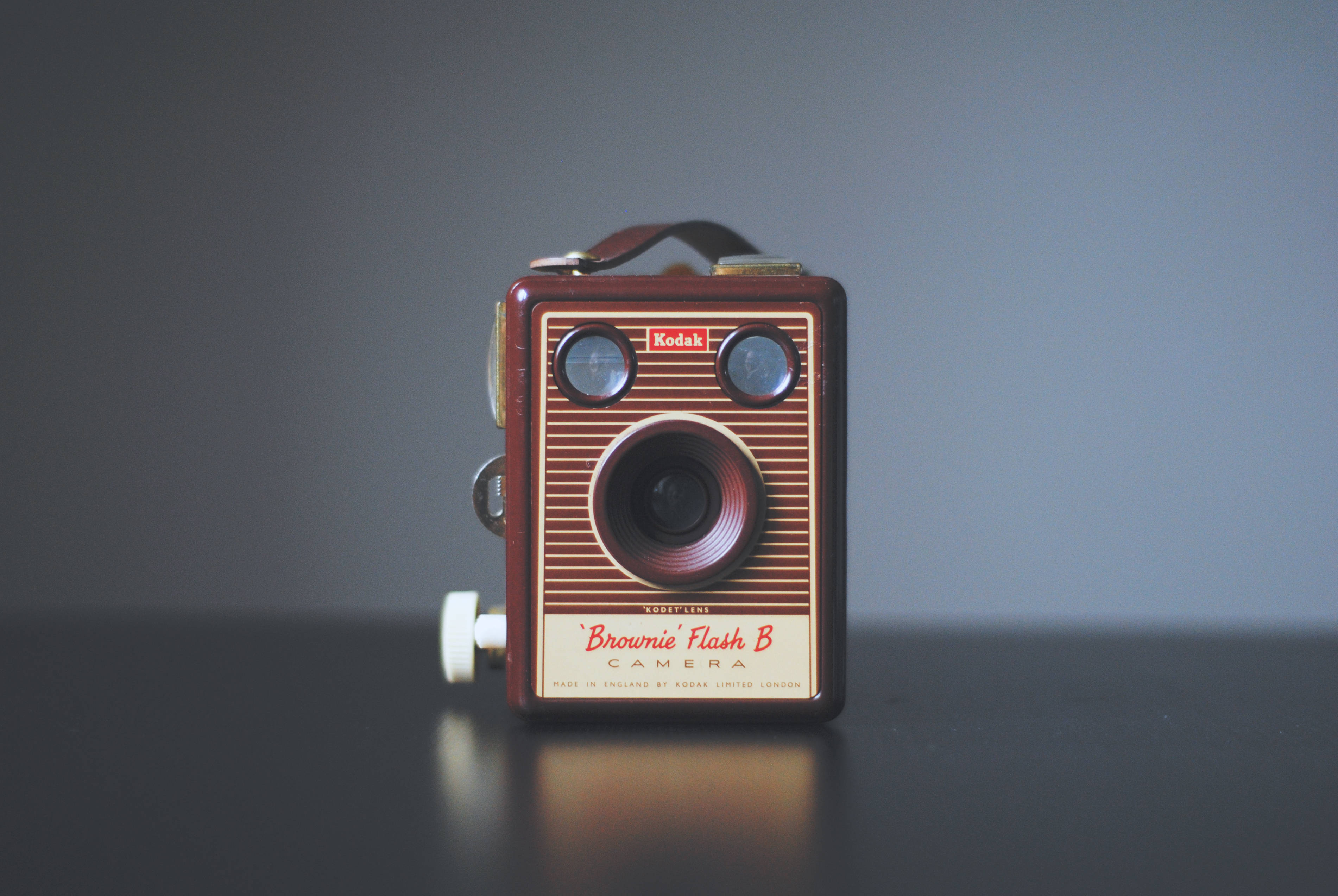 Vintage Minimalist 桌布 壁紙 — 一台復古棕色Kodak Brownie Flash B相機居中放置在深色反光表面上，背景為柔和的灰色。