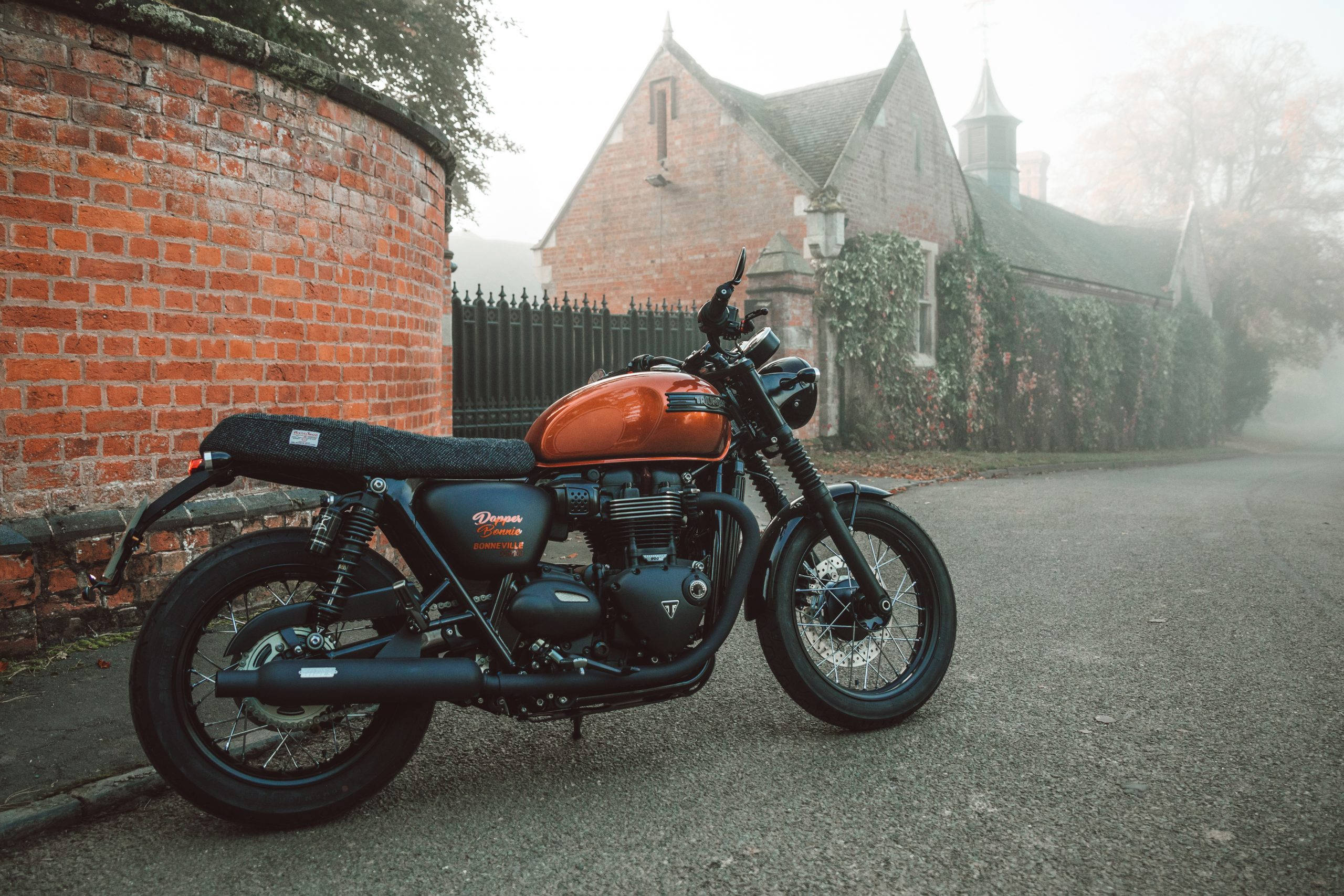 Triumph 桌布 壁紙 — 一台復古橙色和黑色機車停放在霧氣瀰漫的鵝卵石街道上，旁邊是紅磚牆和舊石建築。