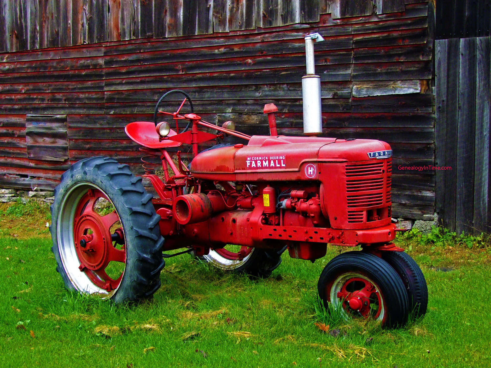 Tractor 桌布 壁紙 — 一台復古紅色 Farmall 拖拉機，配有大黑色輪胎，停在綠色草地上，前面是風化的木製穀倉。