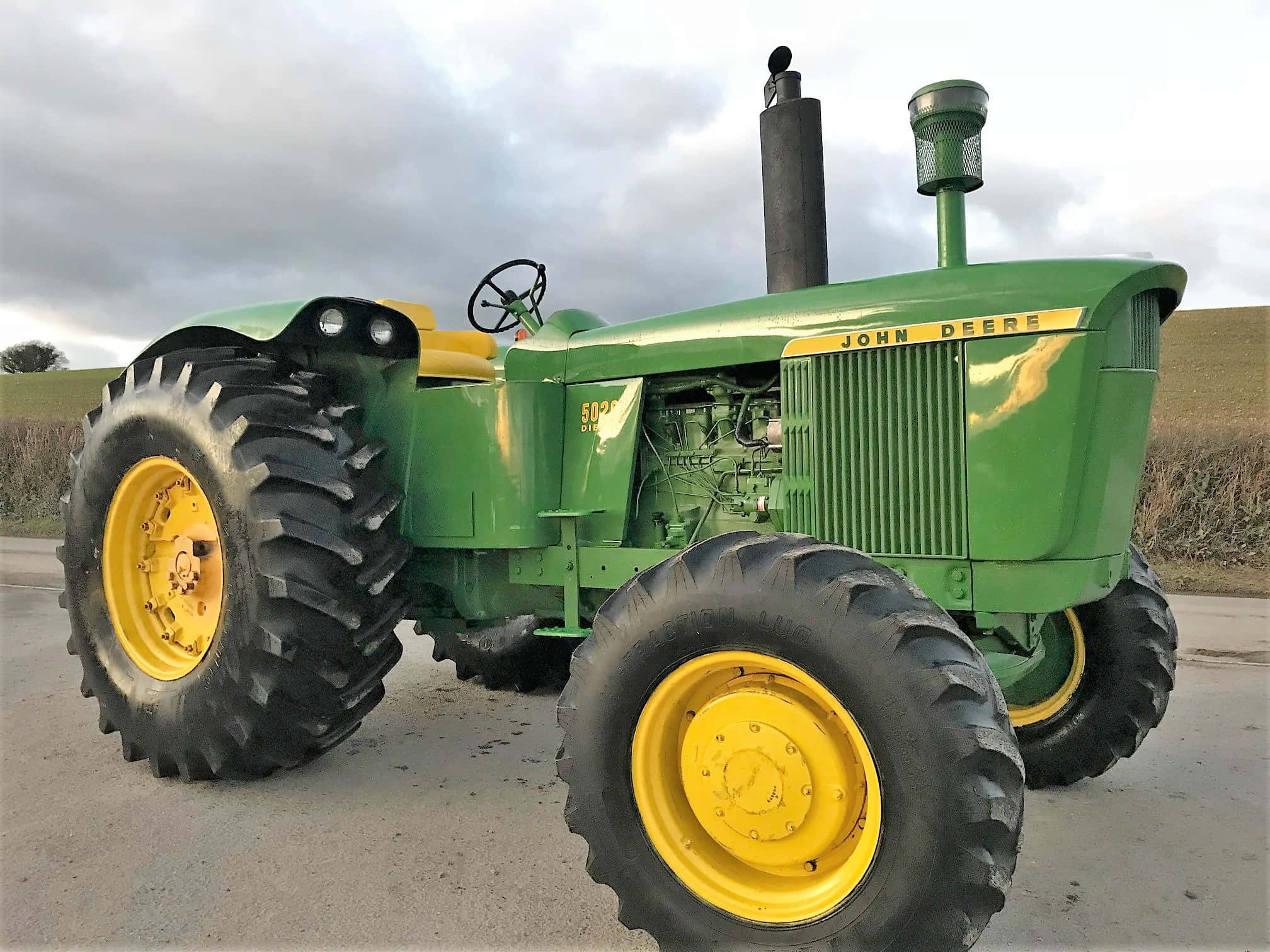 Tractor 桌布 壁紙 — 一台復古綠色和黃色的約翰迪爾拖拉機，配有大黑色輪胎，停在鋪砌的道路上，天空多雲。
