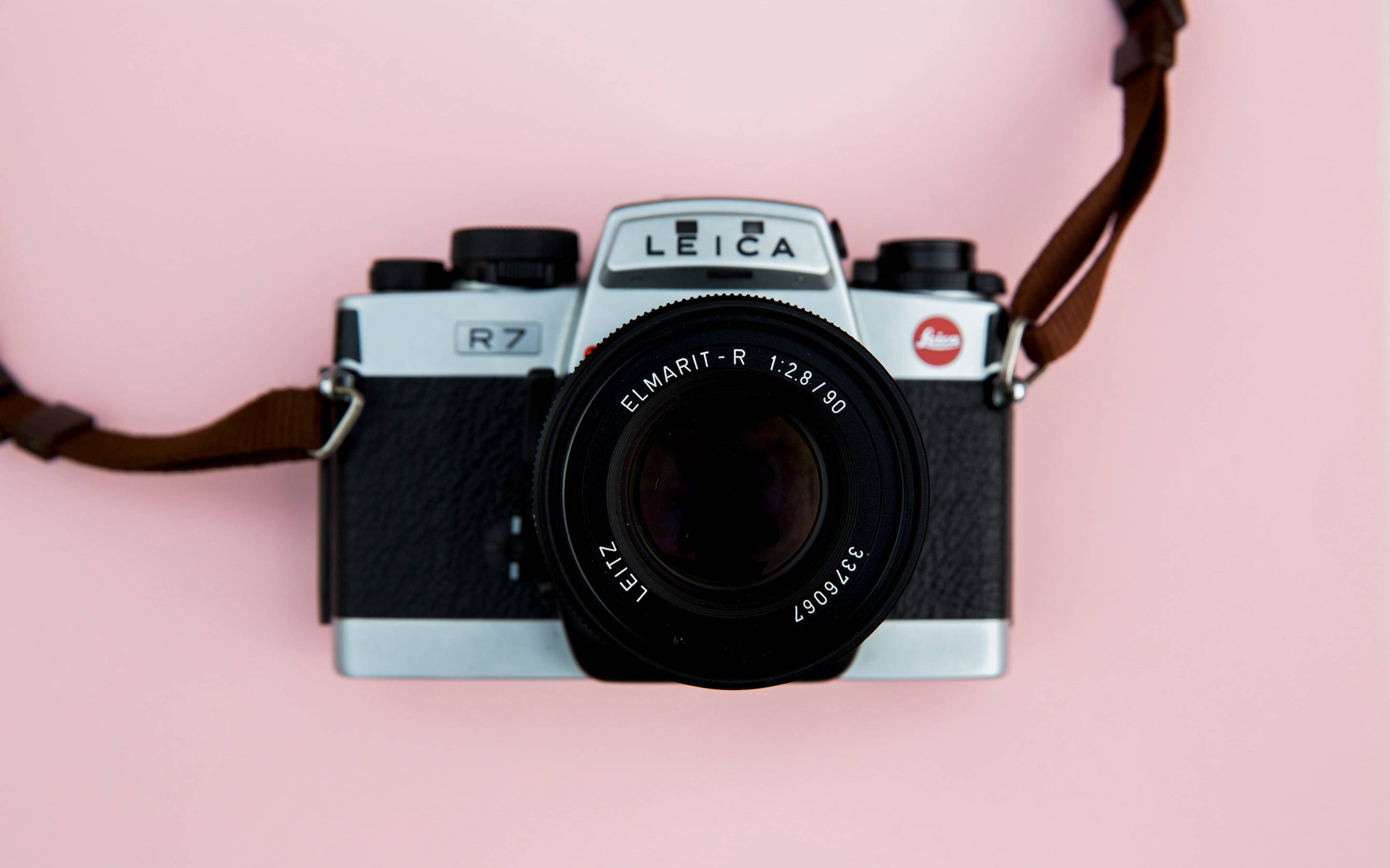 粉紅色 桌布 壁紙 — 一台復古銀色Leica R7底片相機，配有黑色鏡頭和棕色背帶，放置在柔和的粉紅色背景上。