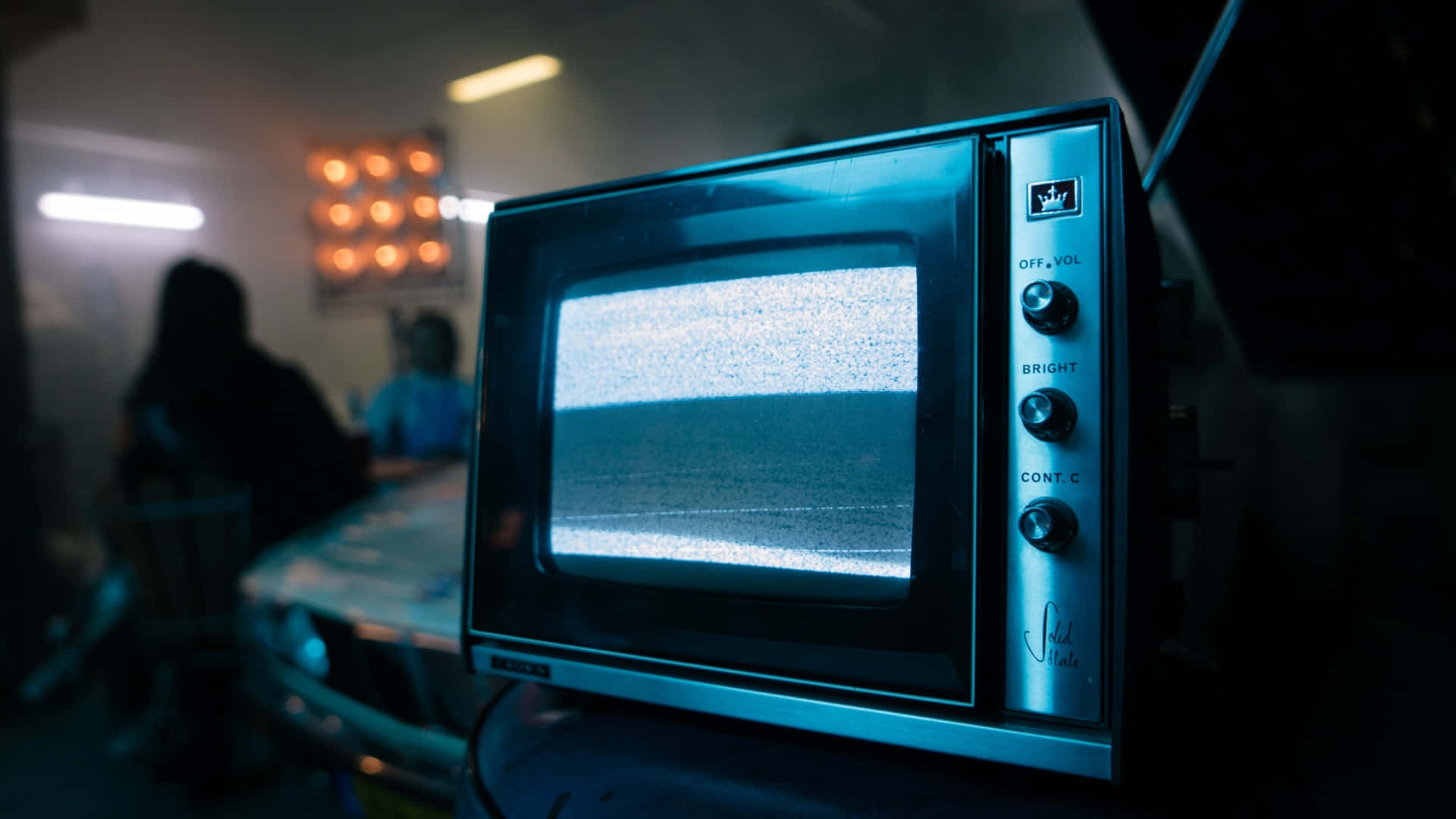 Television Set 桌布 壁紙 — 一台復古電視機螢幕上顯示著雪花靜態畫面，放置在光線昏暗的房間中，背景有模糊的人影。