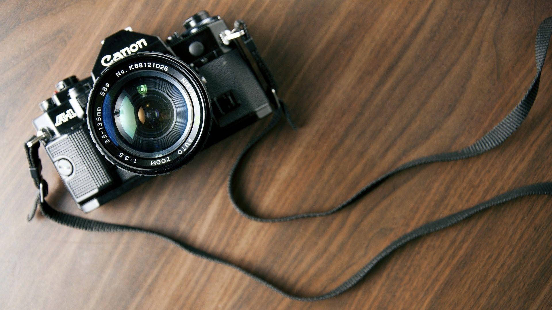 Dslr Camera 桌布 壁紙 — 一台復古黑色佳能相機配備變焦鏡頭，放置在溫暖的木質表面上，相機帶垂掛在附近。