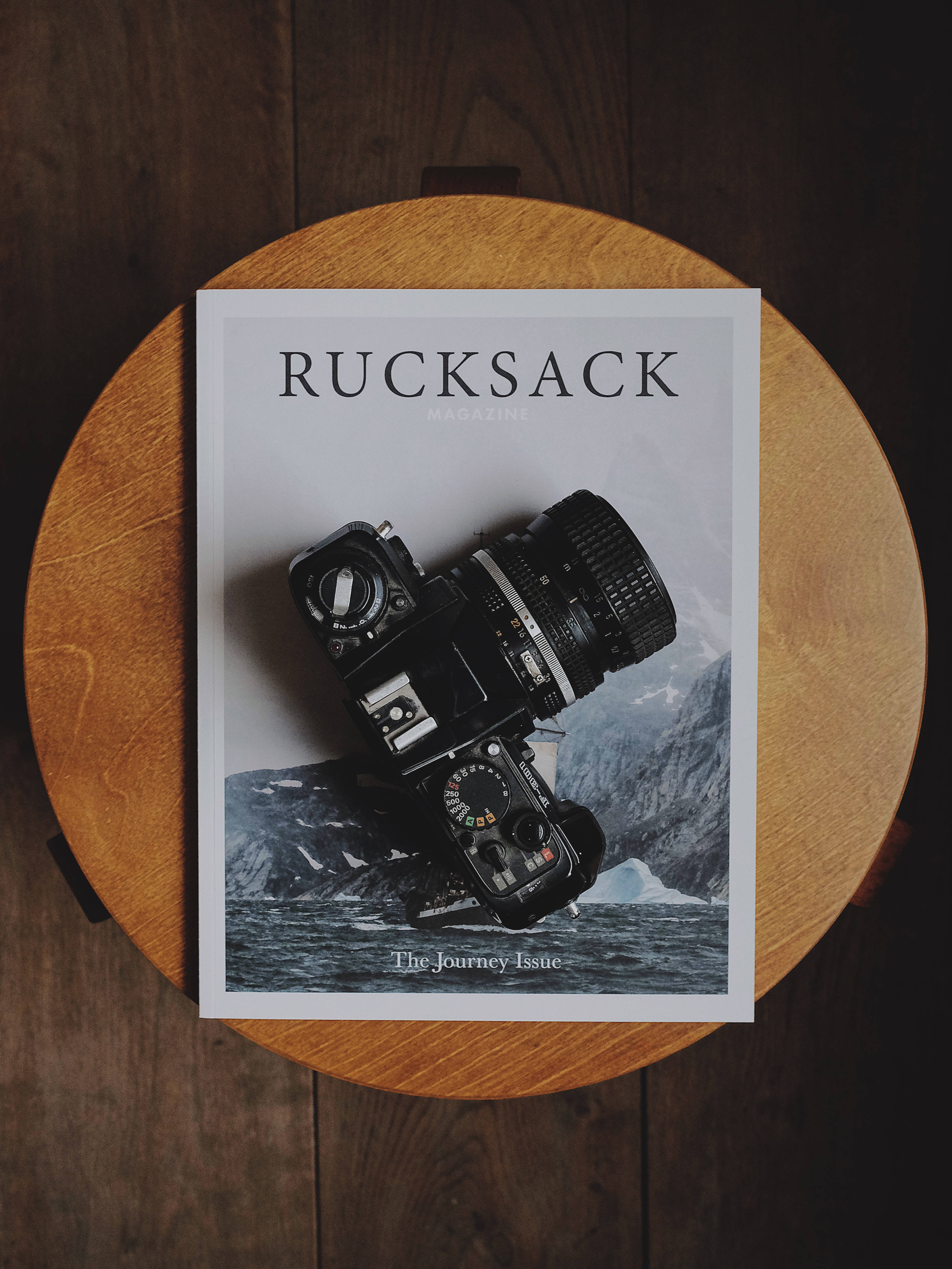 Surface Book 桌布 壁紙 — 一台復古黑色相機放在一本名為「Rucksack」的雜誌上，雜誌置於圓形木桌上，背景為深色木紋。