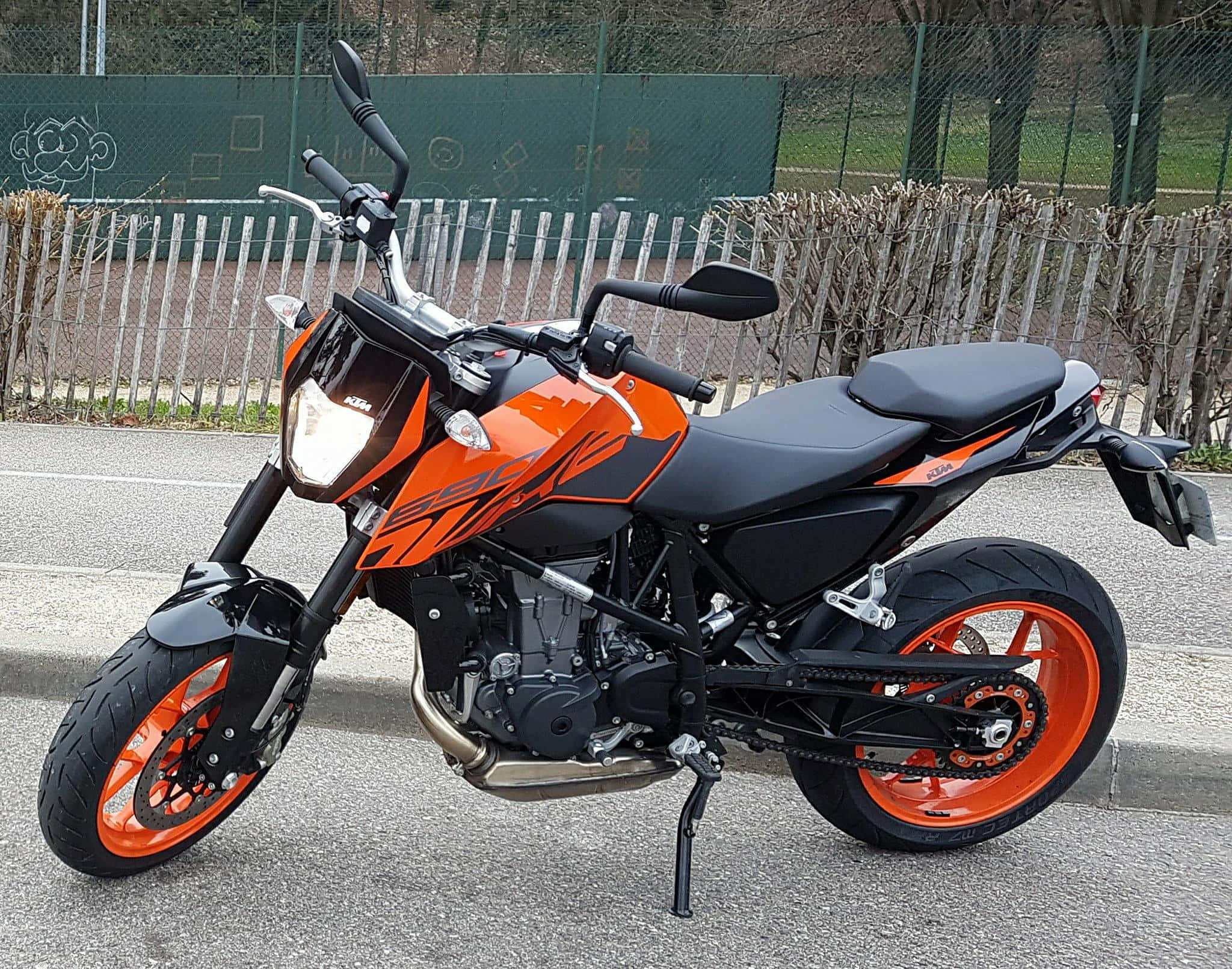 機車 桌布 壁紙 — 一台搶眼的橙色和黑色KTM 390機車停放在鋪砌路面上，背景有圍欄和光禿禿的樹木。