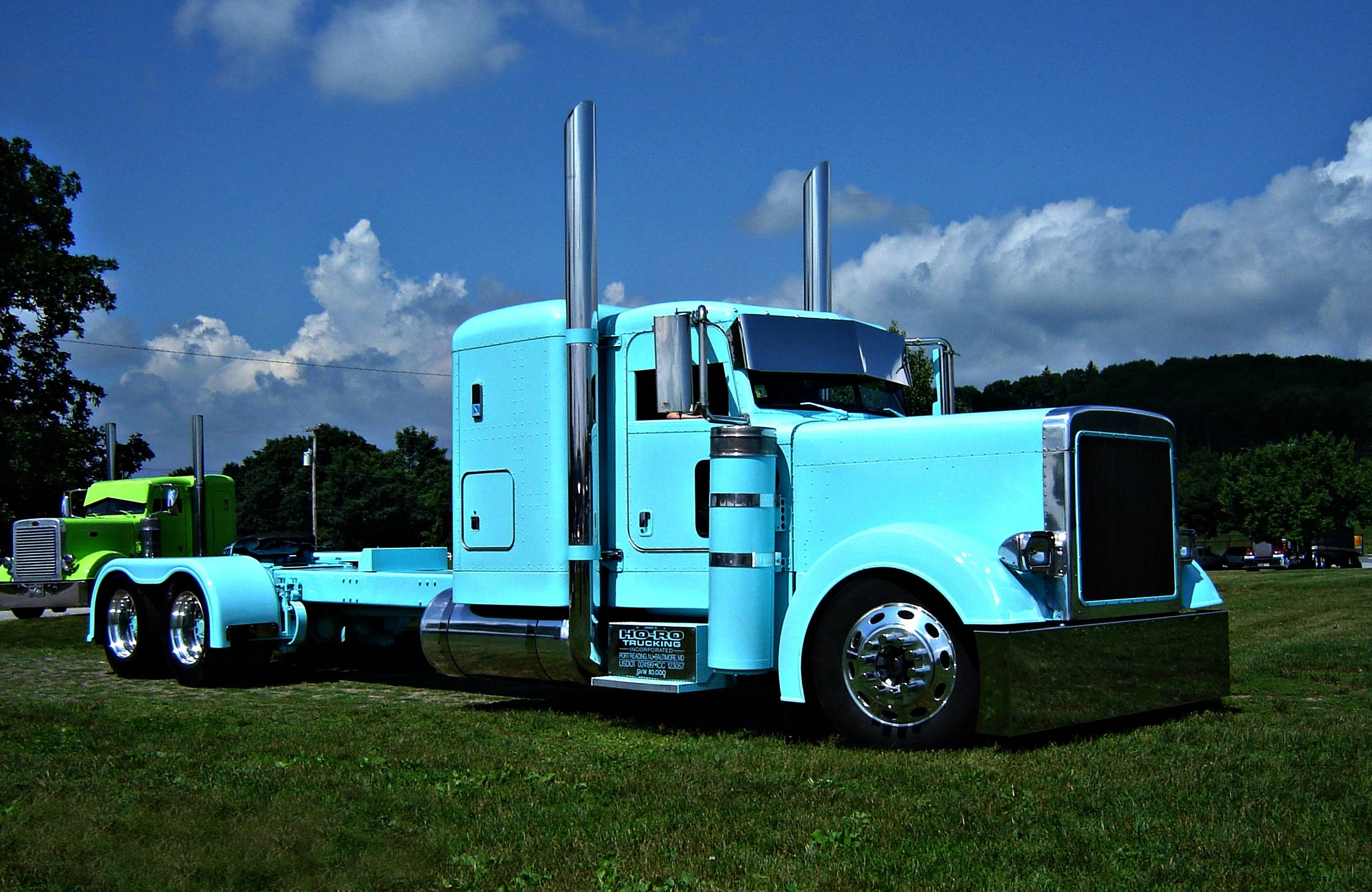 Peterbilt 桌布 壁紙 — 一台搶眼的淺藍色半掛卡車，帶有鍍鉻裝飾，停在草地上，背景是明亮的藍天和蓬鬆的白雲。