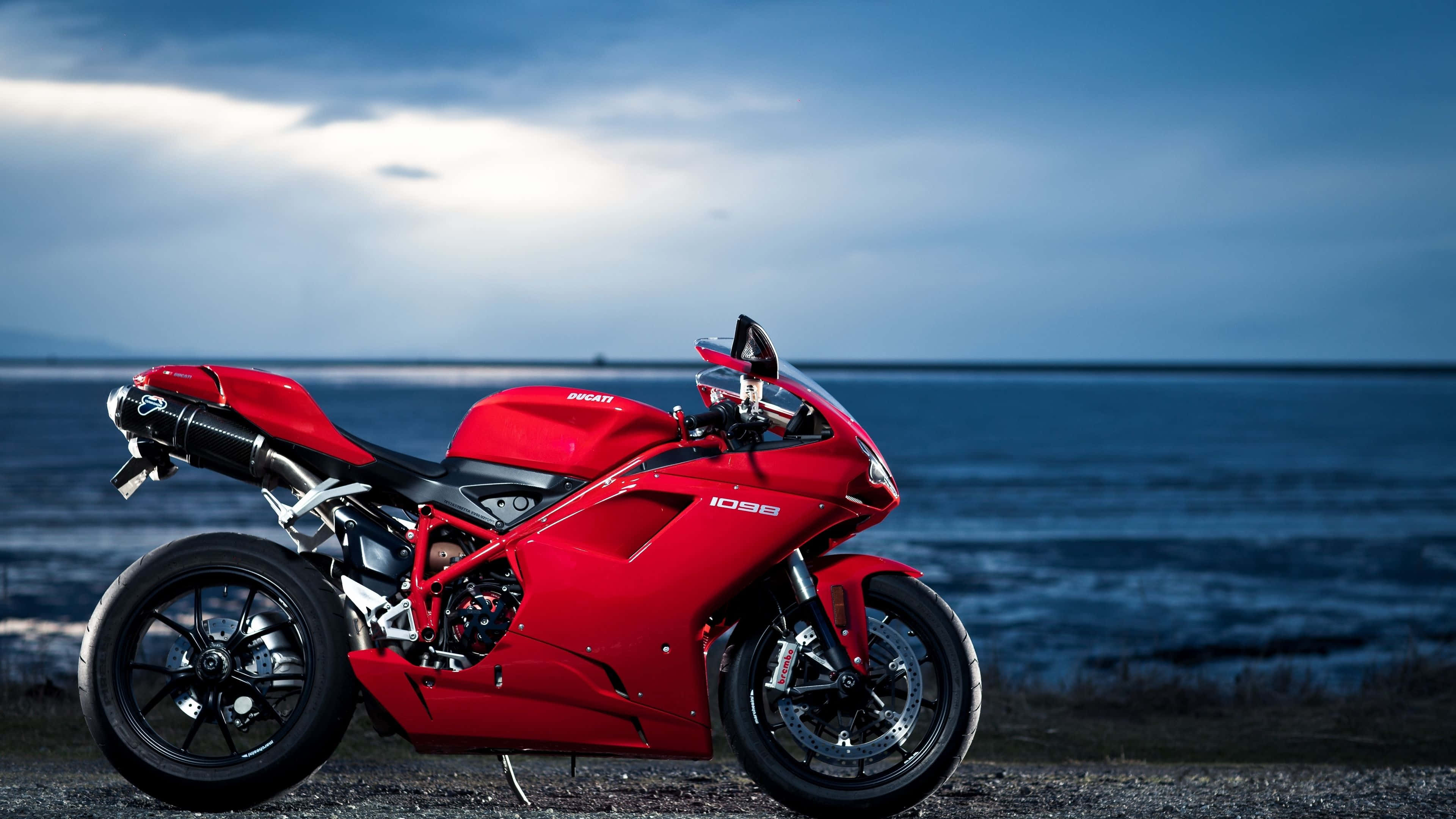 4K Bikes 桌布 壁紙 — 一台搶眼的紅色Ducati 1098機車停在礫石岸邊，背景是廣闊的藍色海洋和陰鬱的黃昏天空。