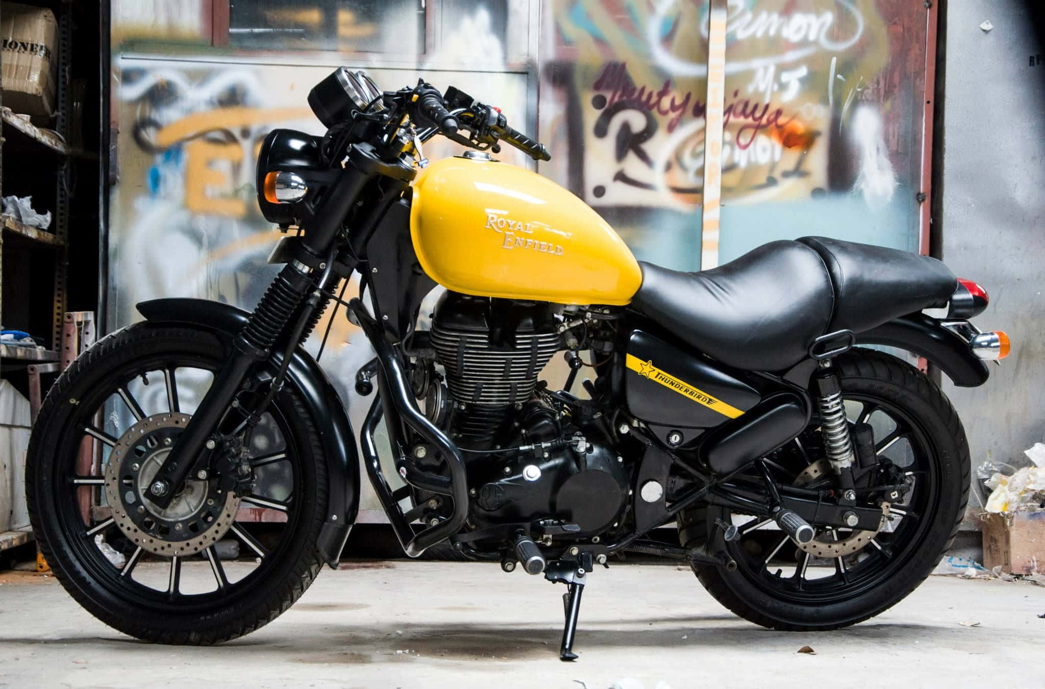 機車 桌布 壁紙 — 一台搶眼的黃黑色 Royal Enfield Thunderbird 摩托車停放在佈滿塗鴉的混凝土車庫內。