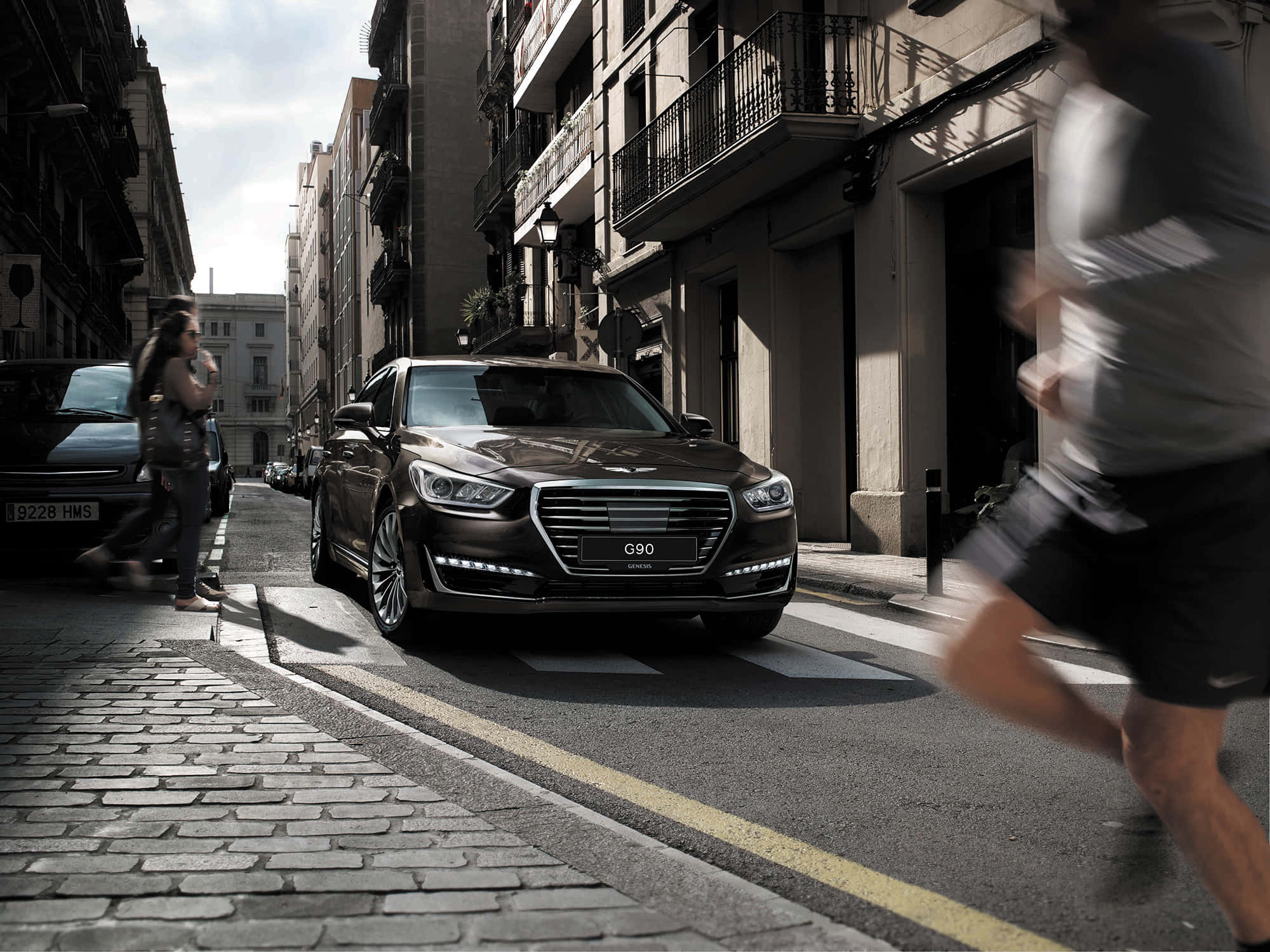 Genesis G90 桌布 壁紙 — 一台時尚深棕色Genesis G90轎車停放在陽光普照的鵝卵石街道上，模糊的行人正在移動。
