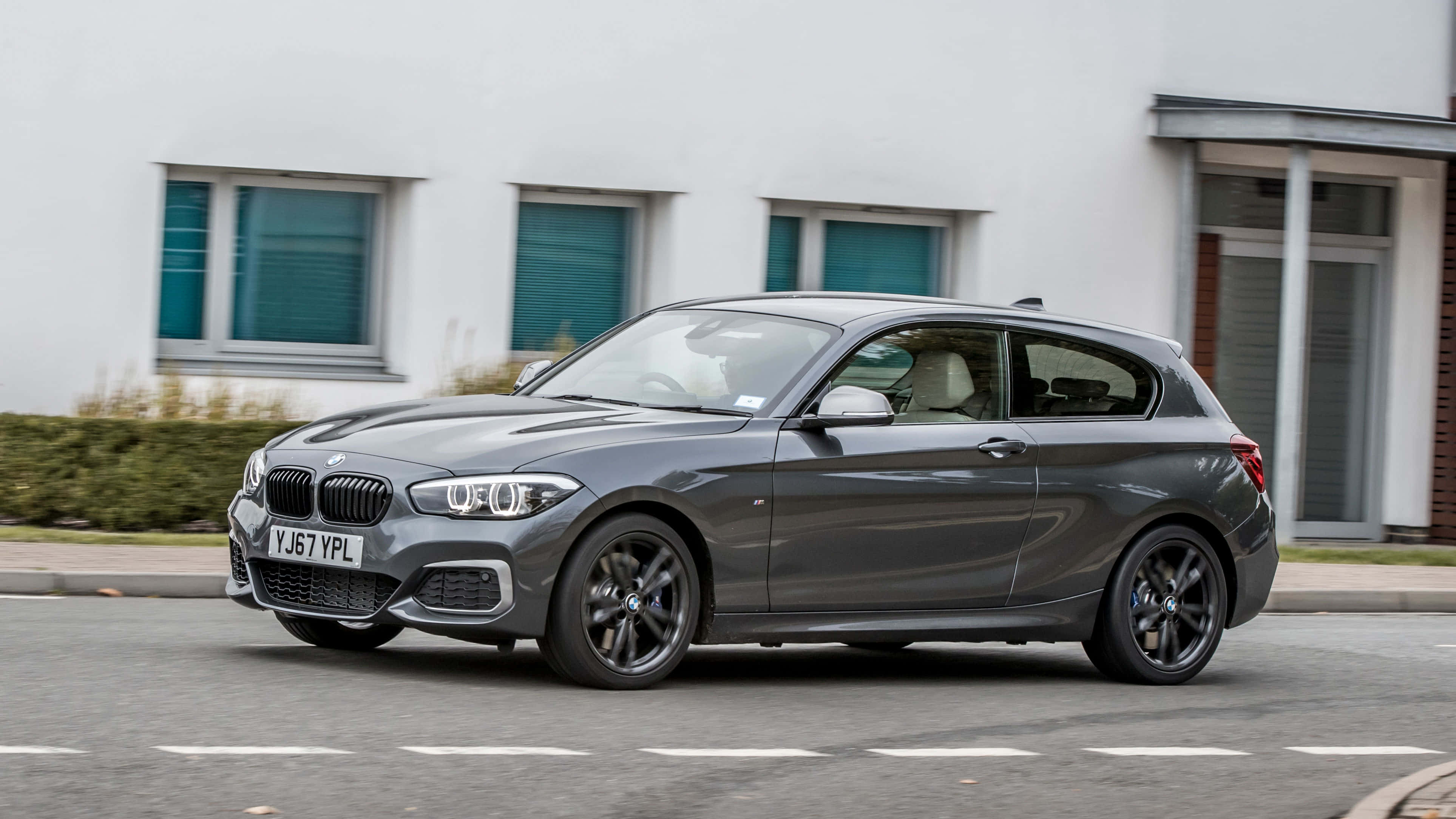 Bmw 1 Series 桌布 壁紙 — 一台時尚灰色BMW 1系列掀背車駛過一棟現代白色建築，建築帶有青綠色窗戶。