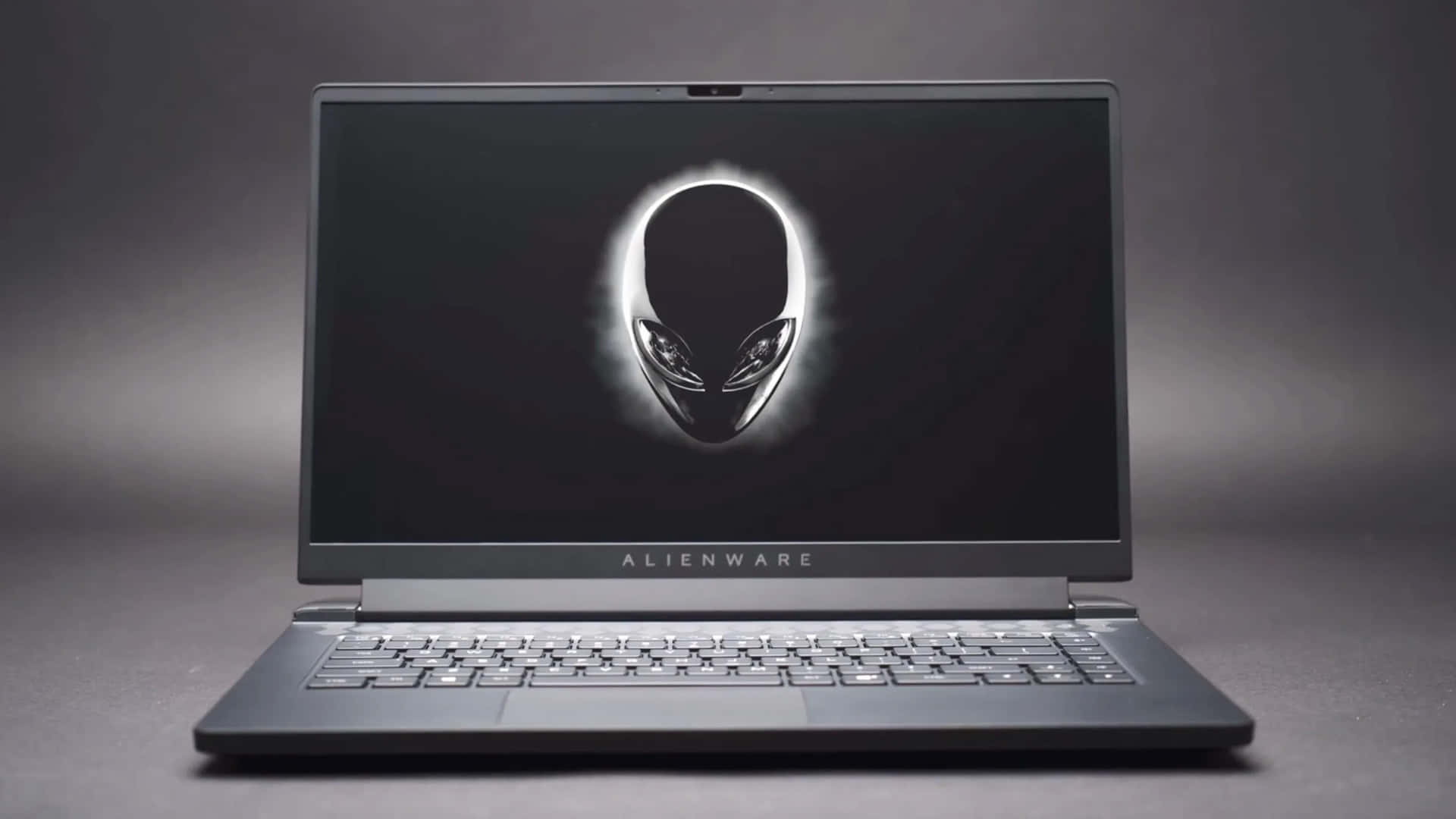 外星人 桌布 壁紙 — 一台時尚的 Alienware 筆記型電腦在深色螢幕上顯示發光的外星人頭標誌，背景為柔和的灰色工作室背景。