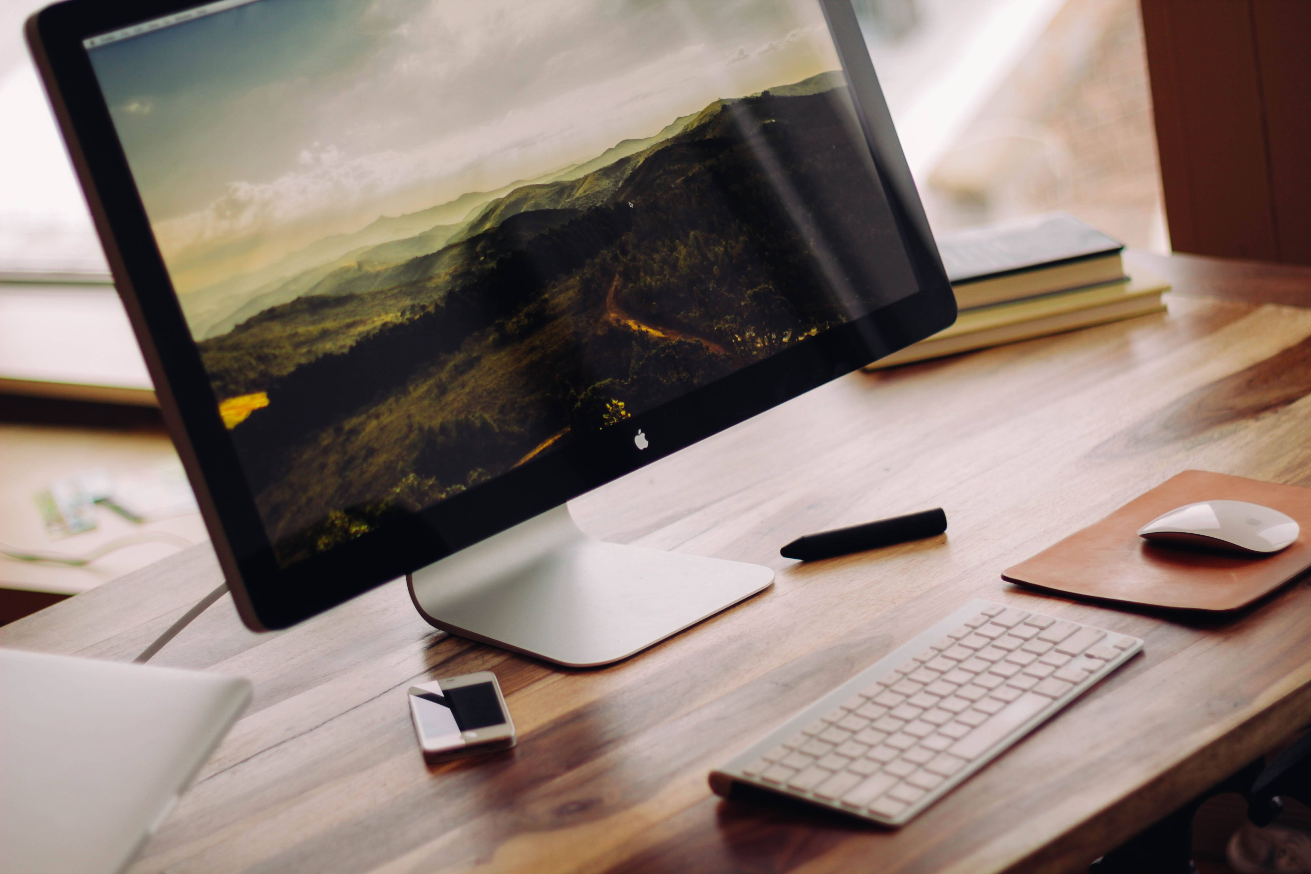 Desk 桌布 壁紙 — 一台時尚的 iMac 放在木桌上，顯示著山景桌布，周圍有鍵盤、滑鼠和電話。