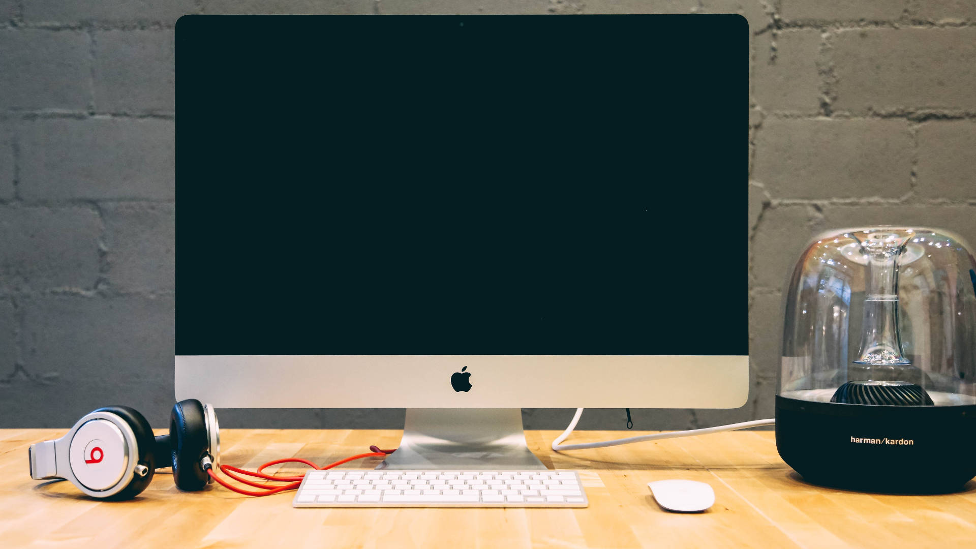 Mac 桌布 壁紙 — 一台時尚的Apple iMac放在木桌上，旁邊有Beats耳機和Harman Kardon喇叭，背景是灰色磚牆。