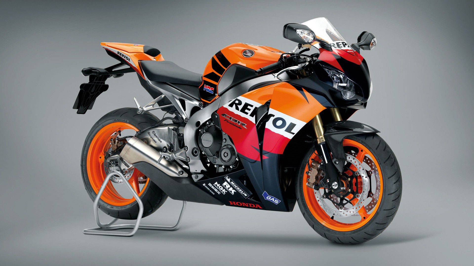 本田 桌布 壁紙 — 一台時尚的Honda Repsol賽車摩托車，帶有鮮豔的橙色和黑色塗裝，在灰色工作室背景前立於支架上。