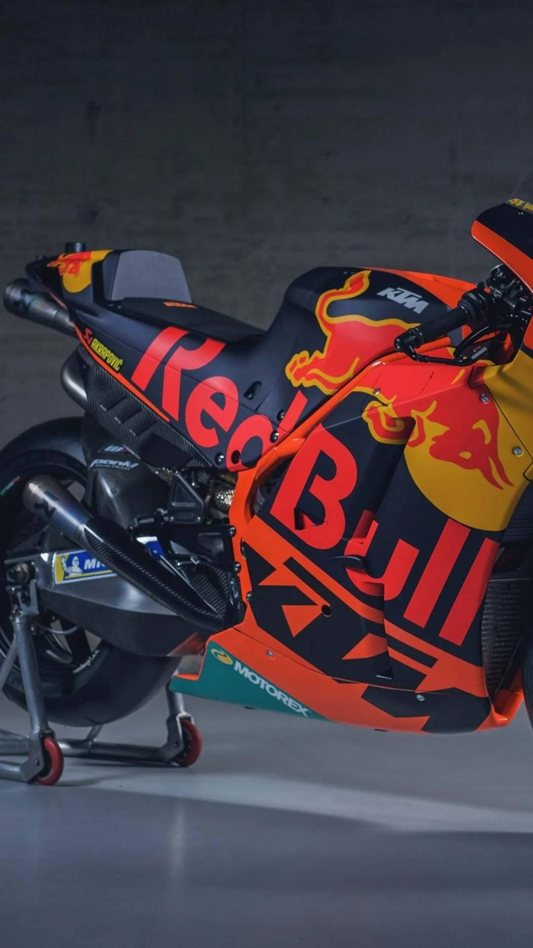  壁紙 — 一台時尚的Red Bull KTM賽車摩托車，帶有醒目的橙色、黑色和黃色塗裝，停在光線昏暗的車庫中的支架上。