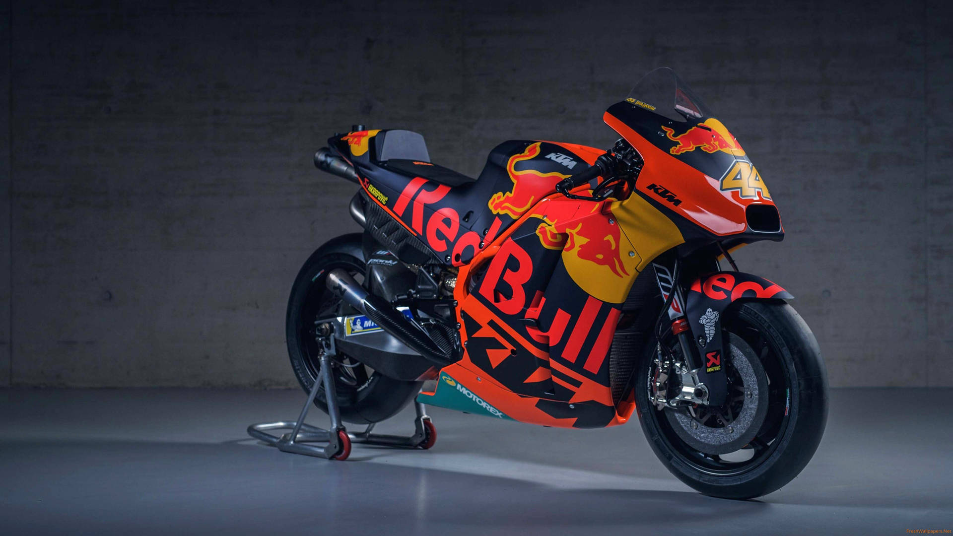  壁紙 — 一台時尚的Red Bull KTM MotoGP賽車，帶有鮮豔的橙色和黑色塗裝，停在支架上，背景是紋理混凝土牆。