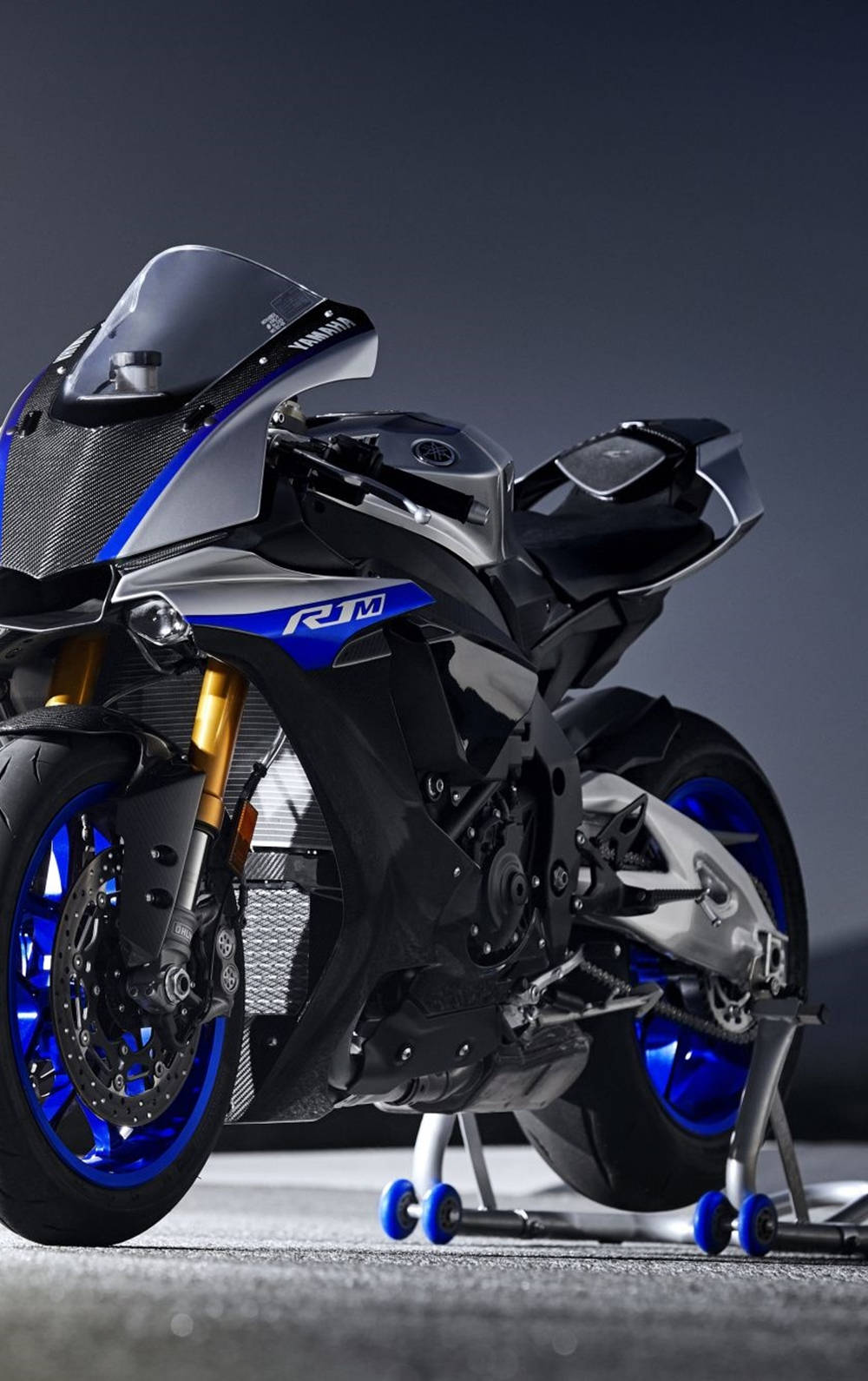 腳踏車 桌布 壁紙 — 一台時尚的Yamaha R1M機車，帶有藍色點綴和金色前叉，在深色攝影棚背景前立於支架上。