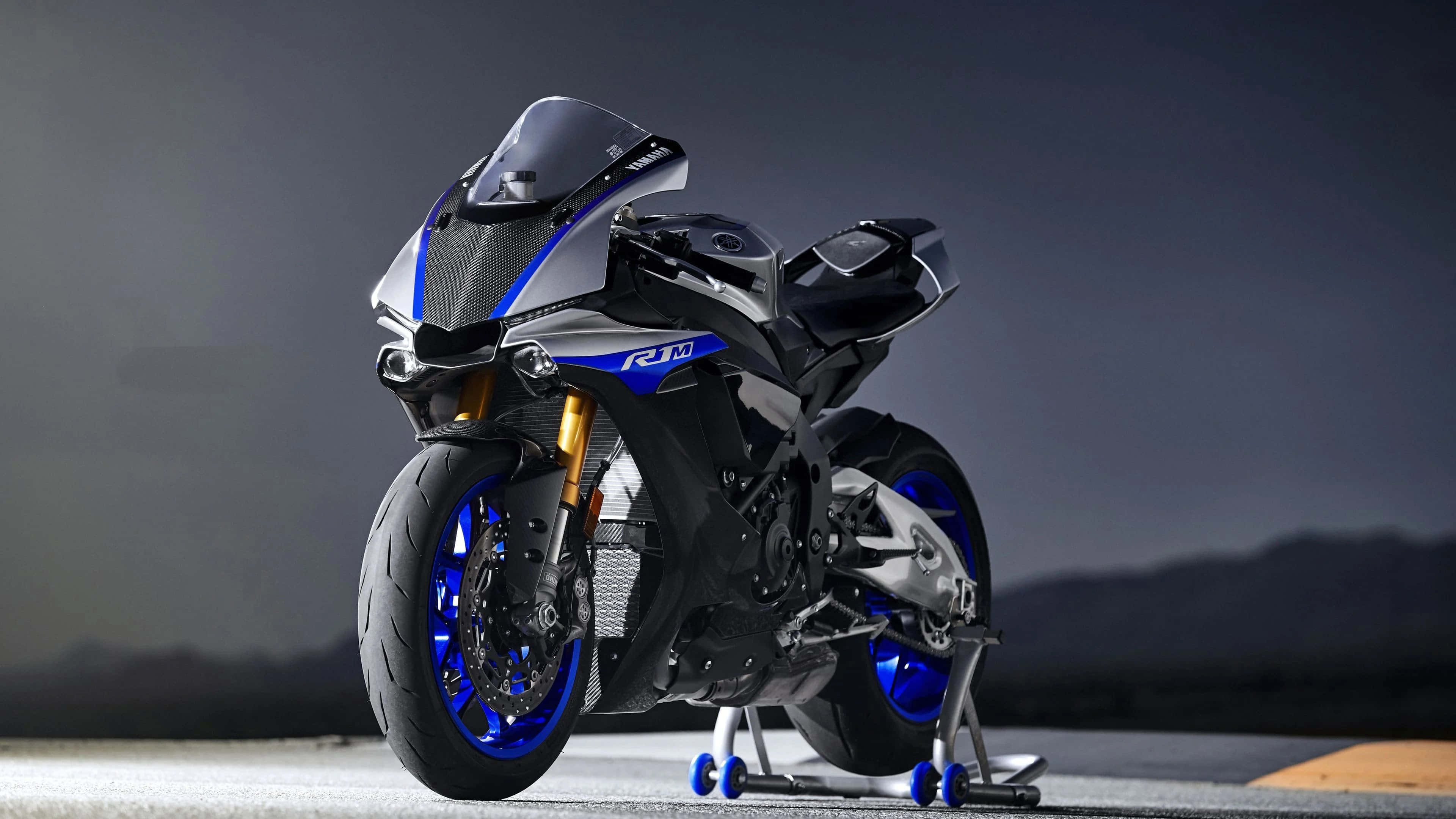 4K Bikes 桌布 壁紙 — 一台時尚的Yamaha R1M運動型摩托車，帶有藍色點綴和金色前叉，在黑暗的山脈背景下蓄勢待發於賽道上。