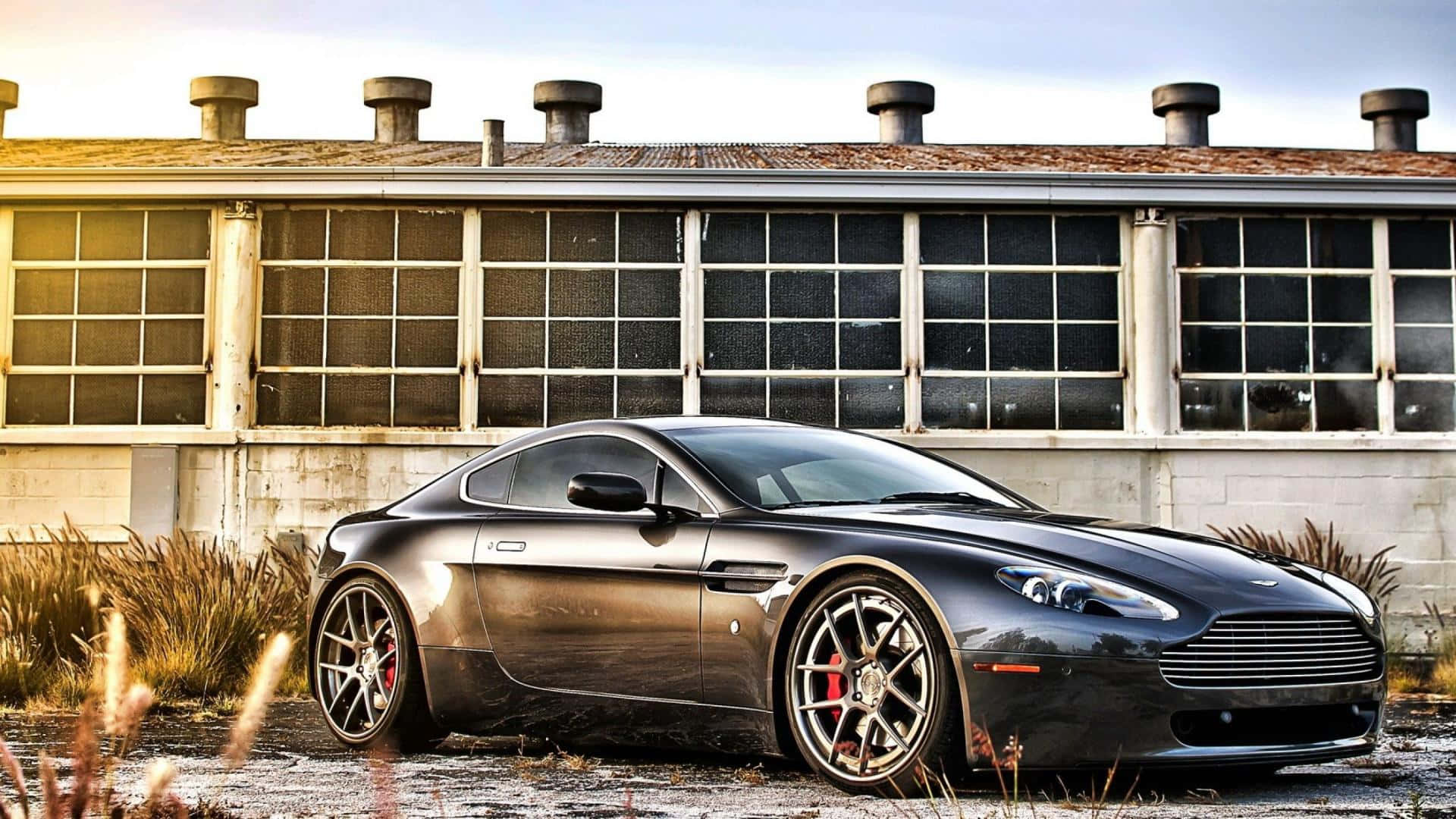 Aston Martin V12 Vantage 桌布 壁紙 — 一台時尚的啞光黑色Aston Martin跑車停放在一個帶有大窗戶、風化了的工業建築前。