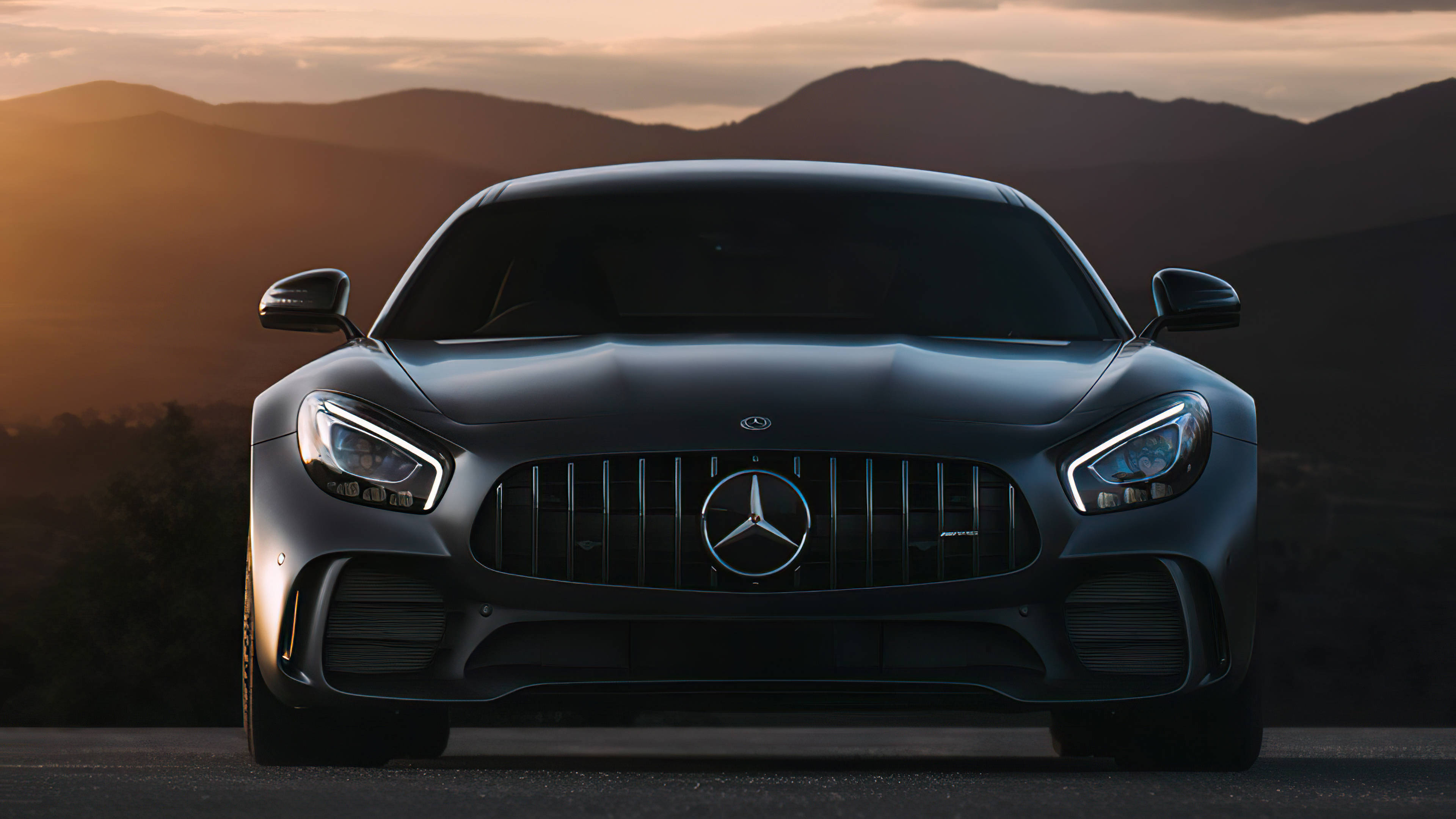 Mercedes Benz Amg 桌布 壁紙 — 一台時尚的啞光黑色Mercedes-AMG GT停在道路上，背後是被金色夕陽沐浴的山脈。