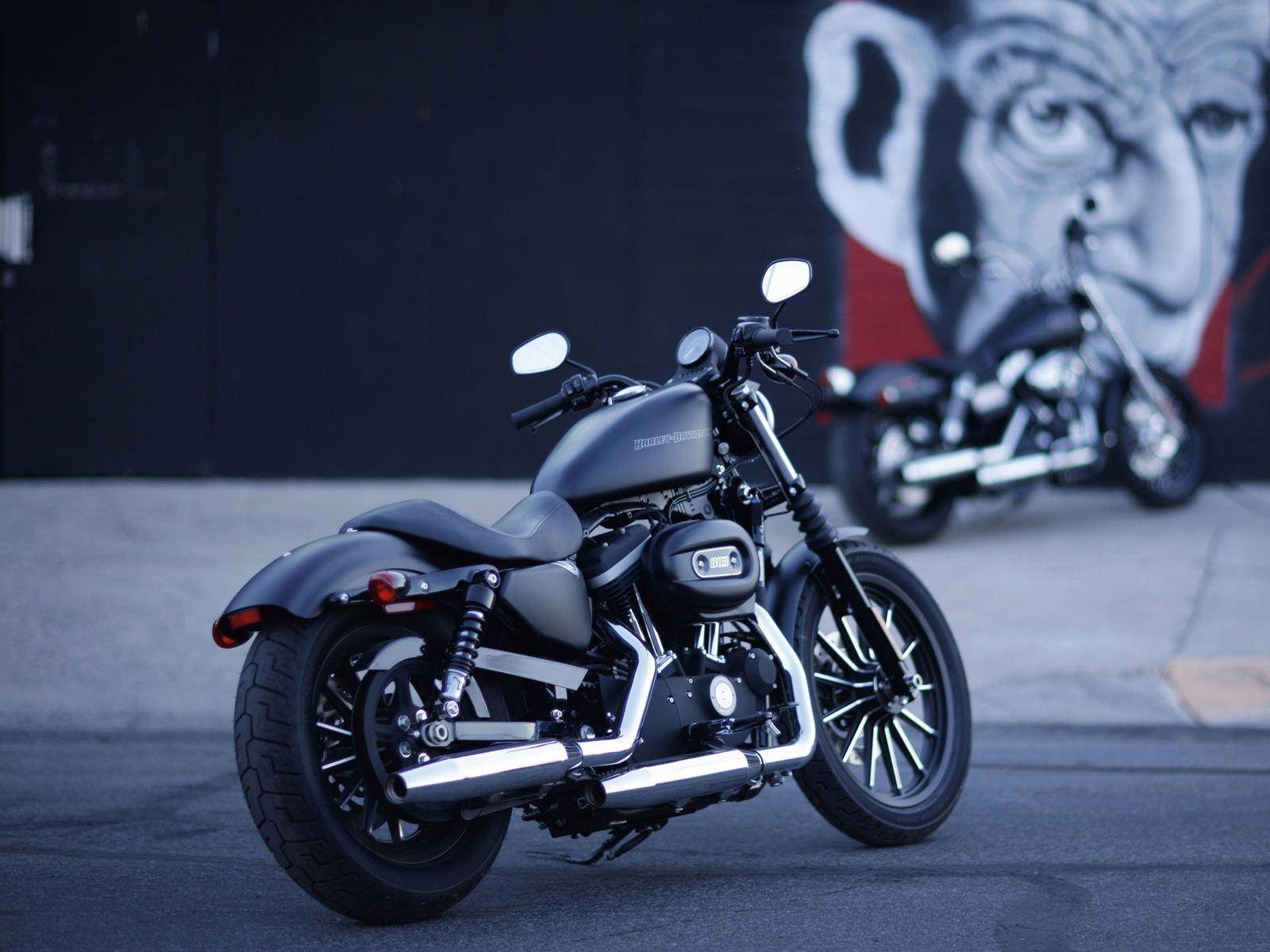 Harley 桌布 壁紙 — 一台時尚的啞光黑色摩托車停在人行道上，背景是一幅大型的塗鴉壁畫，畫著一張臉。