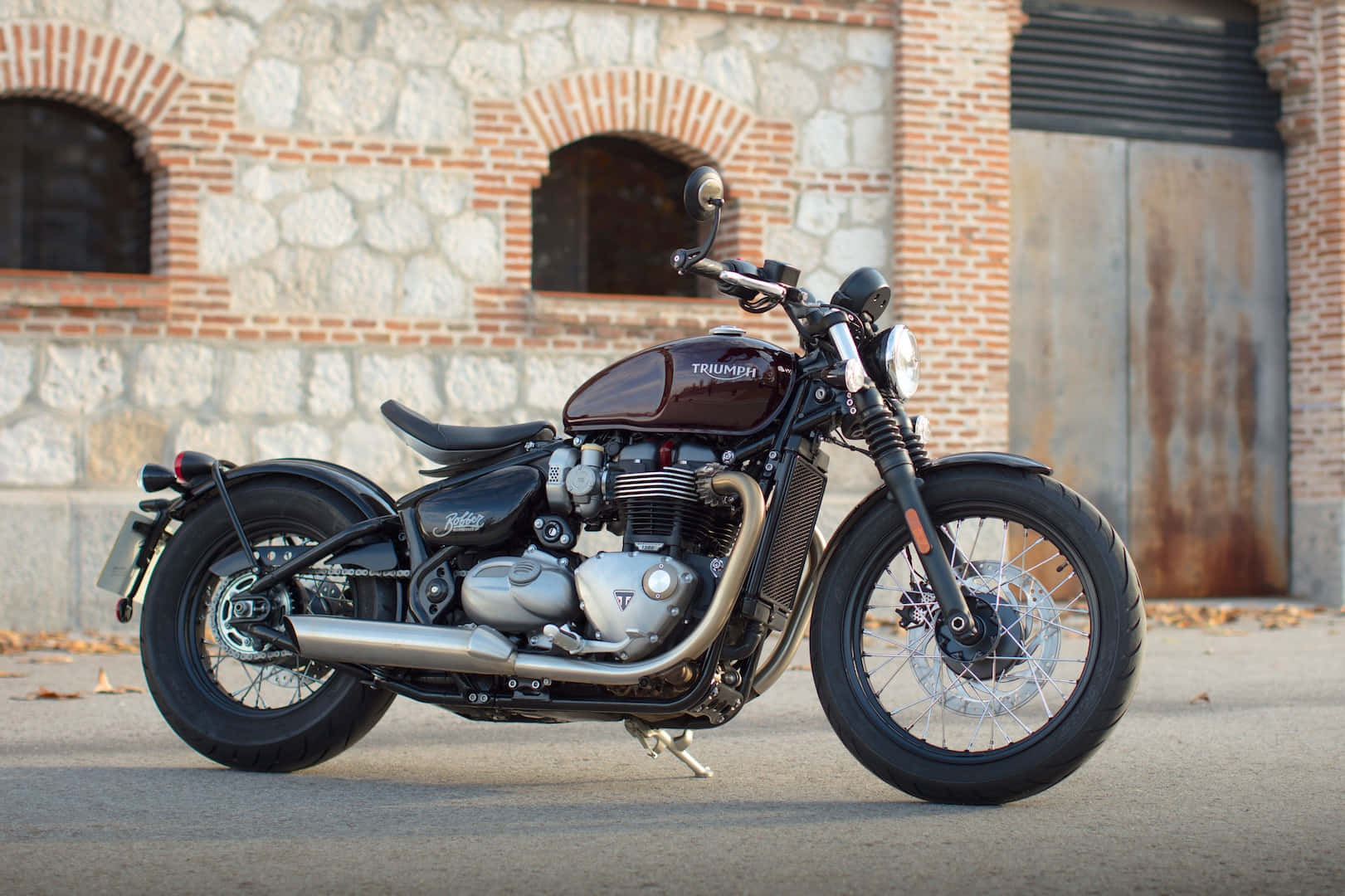 機車 桌布 壁紙 — 一台時尚的栗色 Triumph 摩托車停放在鋪砌的街道上，前面是帶有拱形窗戶的質樸磚石建築。
