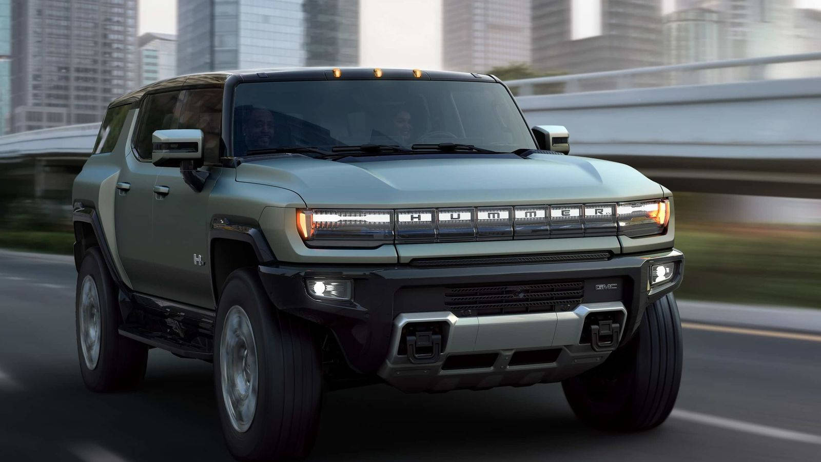 Truck 桌布 壁紙 — 一台時尚的橄欖綠色Hummer EV SUV在模糊的城市高速公路上行駛，背景是高樓大廈。