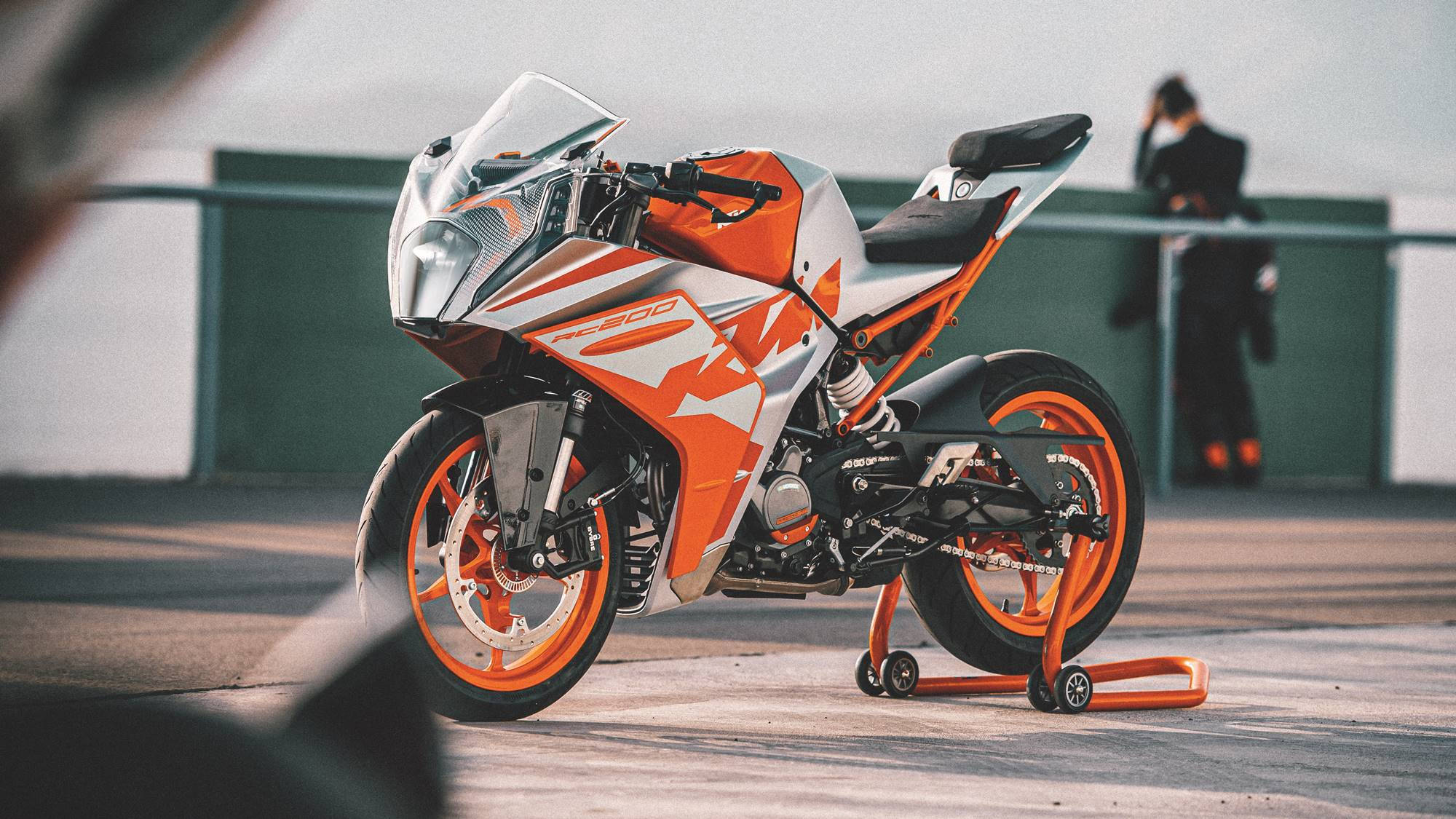 KTM RC 200 桌布 壁紙 — 一台時尚的橙色和白色KTM RC200運動機車放在混凝土平台上的支架上，背景有一個模糊的人影。
