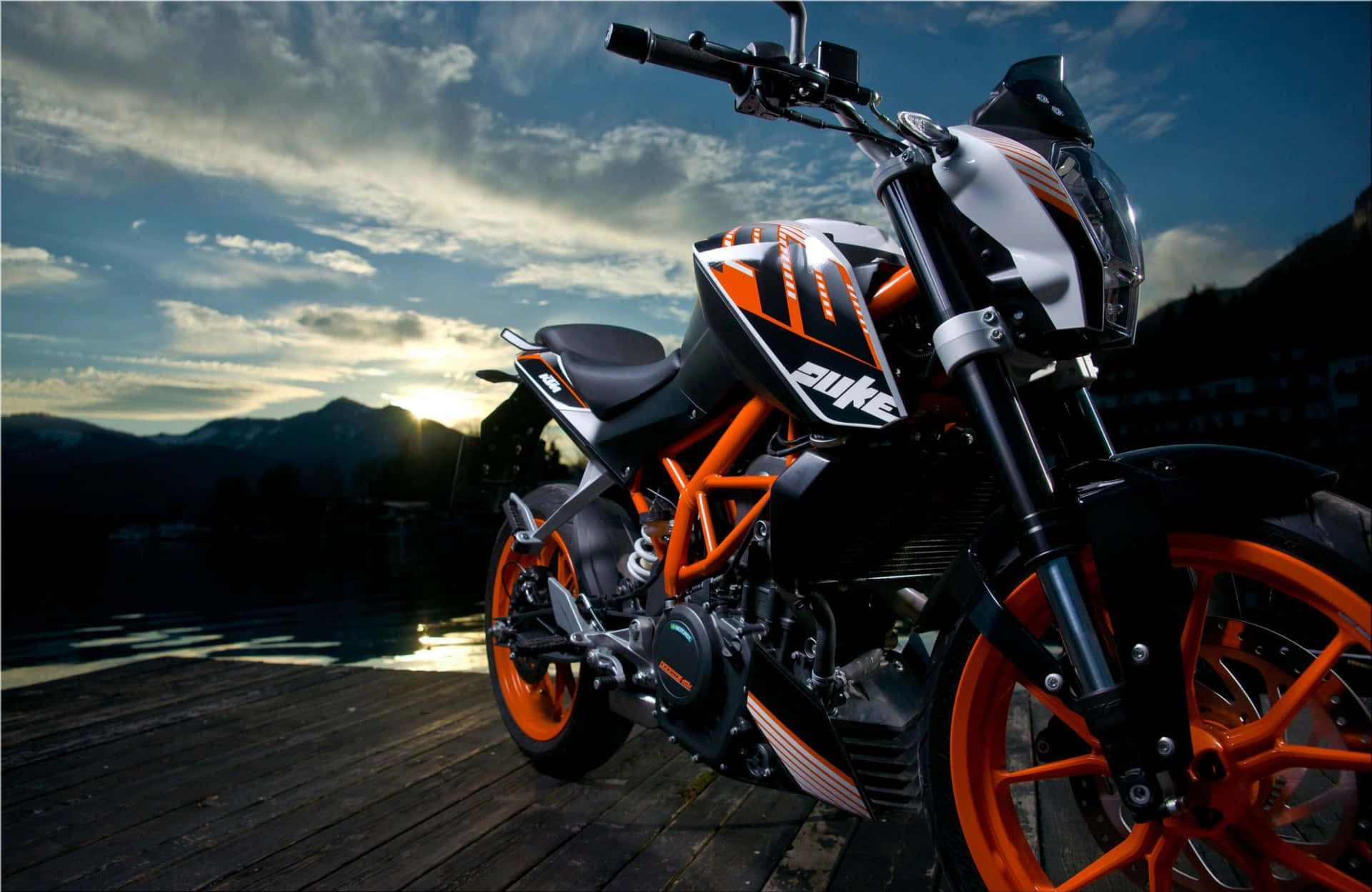  壁紙 — 一台時尚的橙色和黑色KTM Duke摩托車停泊在日落時分的木製碼頭上，背景有山脈。