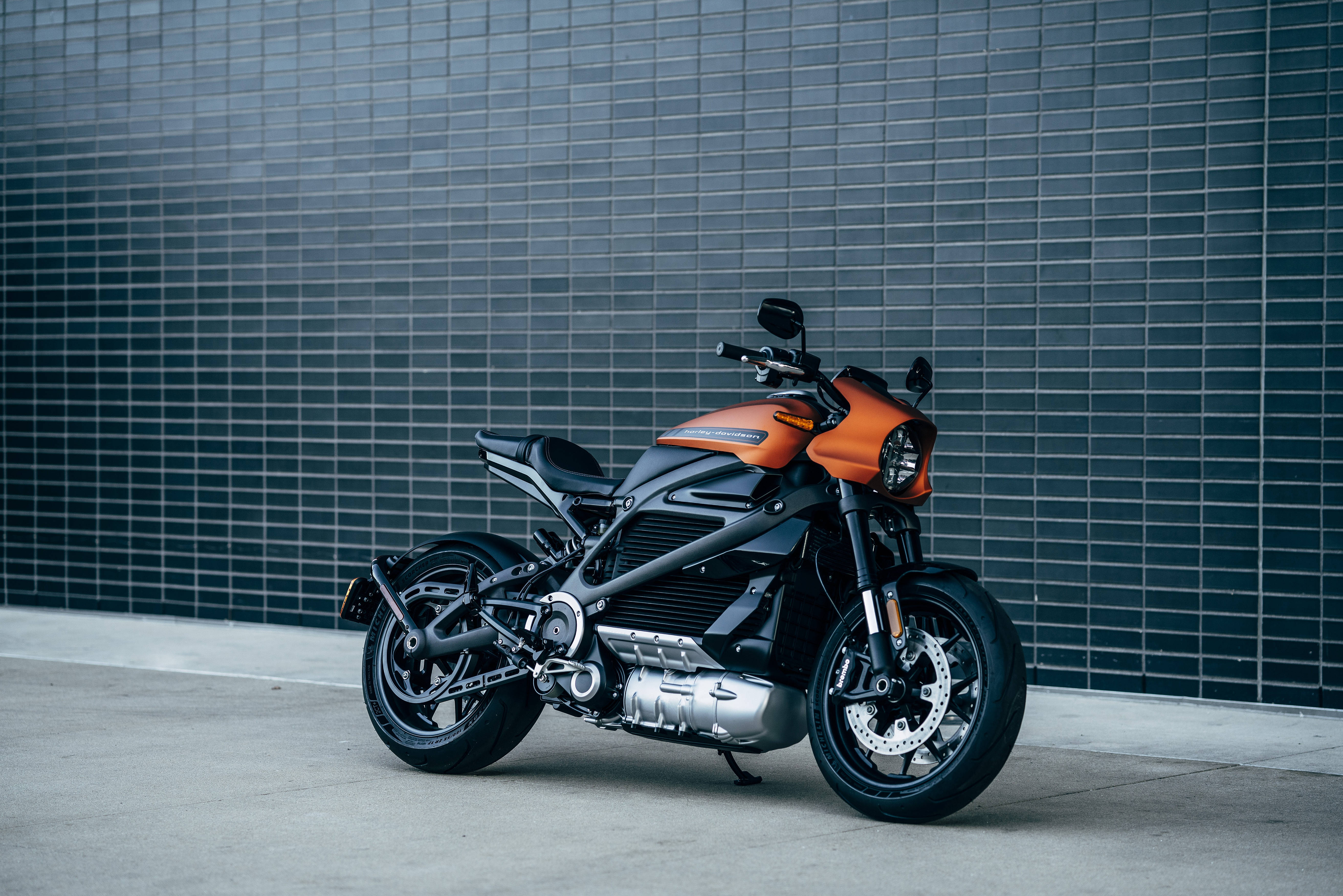 Harley 桌布 壁紙 — 一台時尚的橙色和黑色電動機車，在混凝土表面上靠著深色瓷磚牆矗立。
