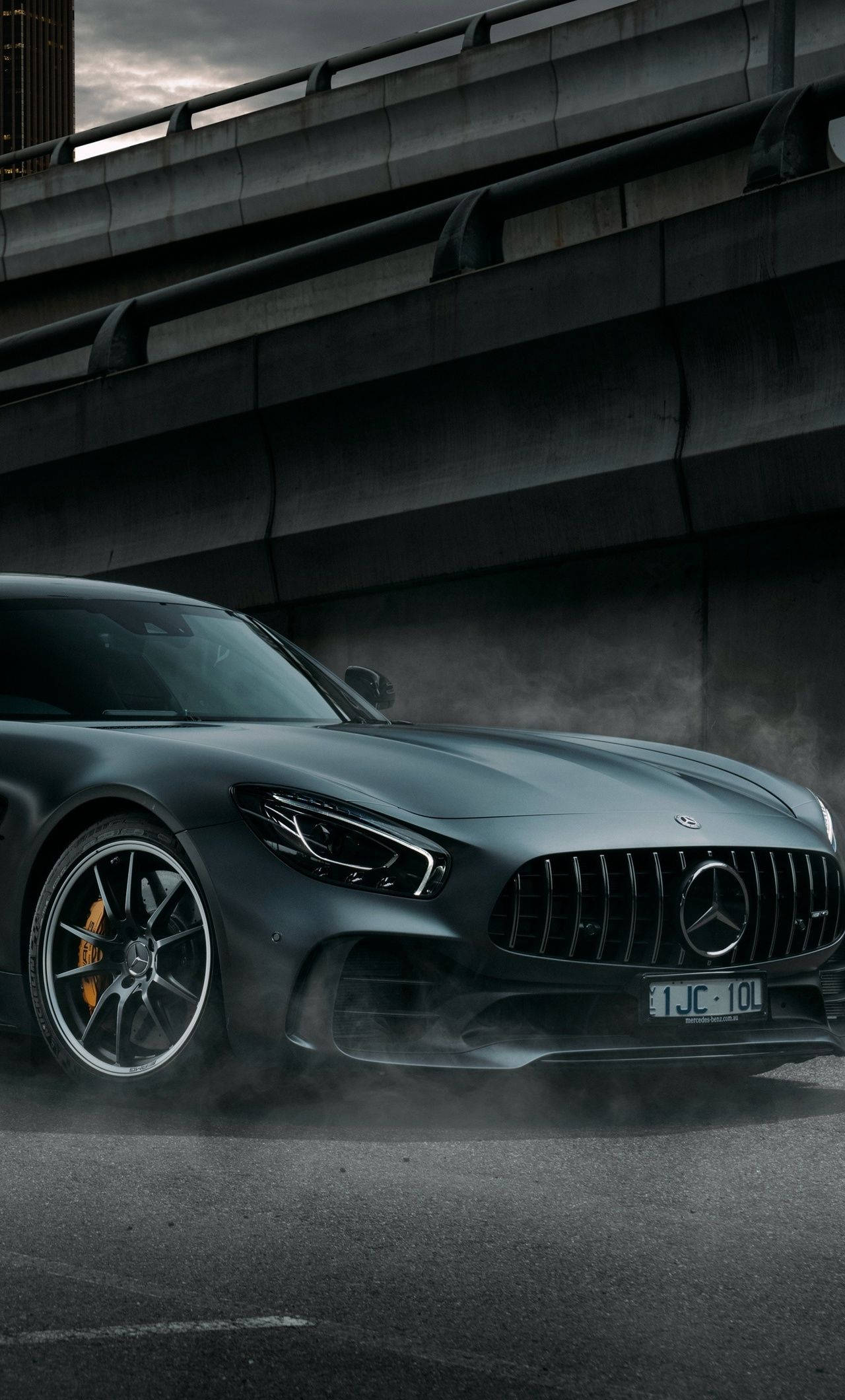 賓士 iPhone 桌布 壁紙 — 一台時尚的消光灰色 Mercedes-AMG GT 跑車從混凝土天橋下駛出，輪胎周圍煙霧繚繞。