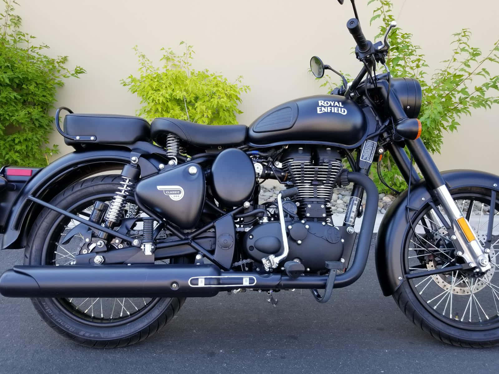 機車 桌布 壁紙 — 一台時尚的消光黑色 Royal Enfield 摩托車停在柏油路上，後面有綠色灌木叢。
