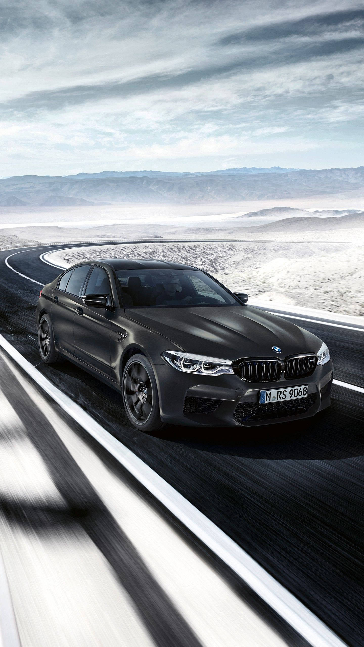 X 桌布 壁紙 — 一台時尚的消光黑色BMW M5 RS在陰天、遠山環繞的蜿蜒沙漠高速公路上奔馳。