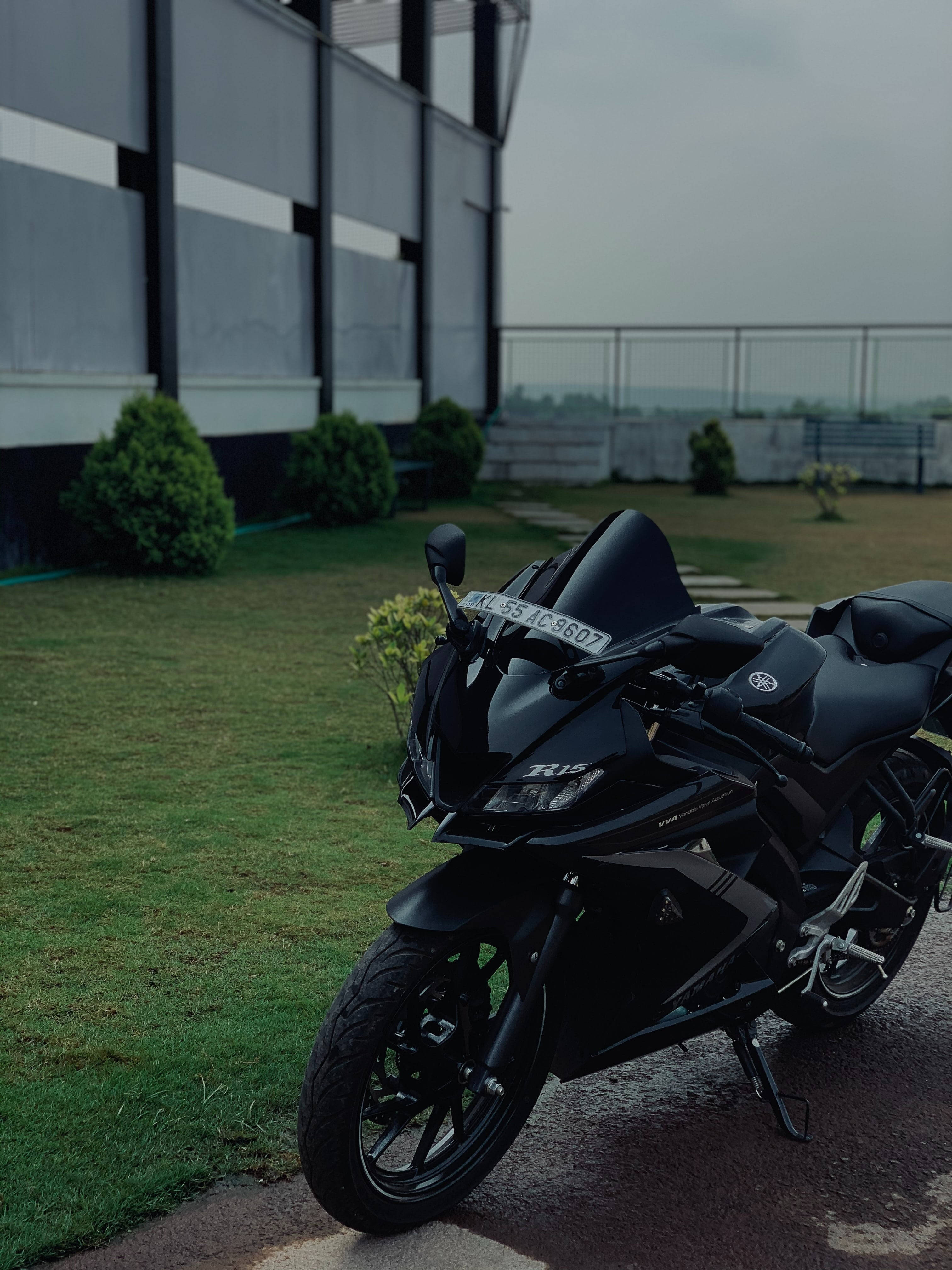桌布 壁紙 — 一台時尚的消光黑色Yamaha R15機車停在濕漉漉的人行道上，旁邊是現代建築物和綠色灌木，天空多雲。