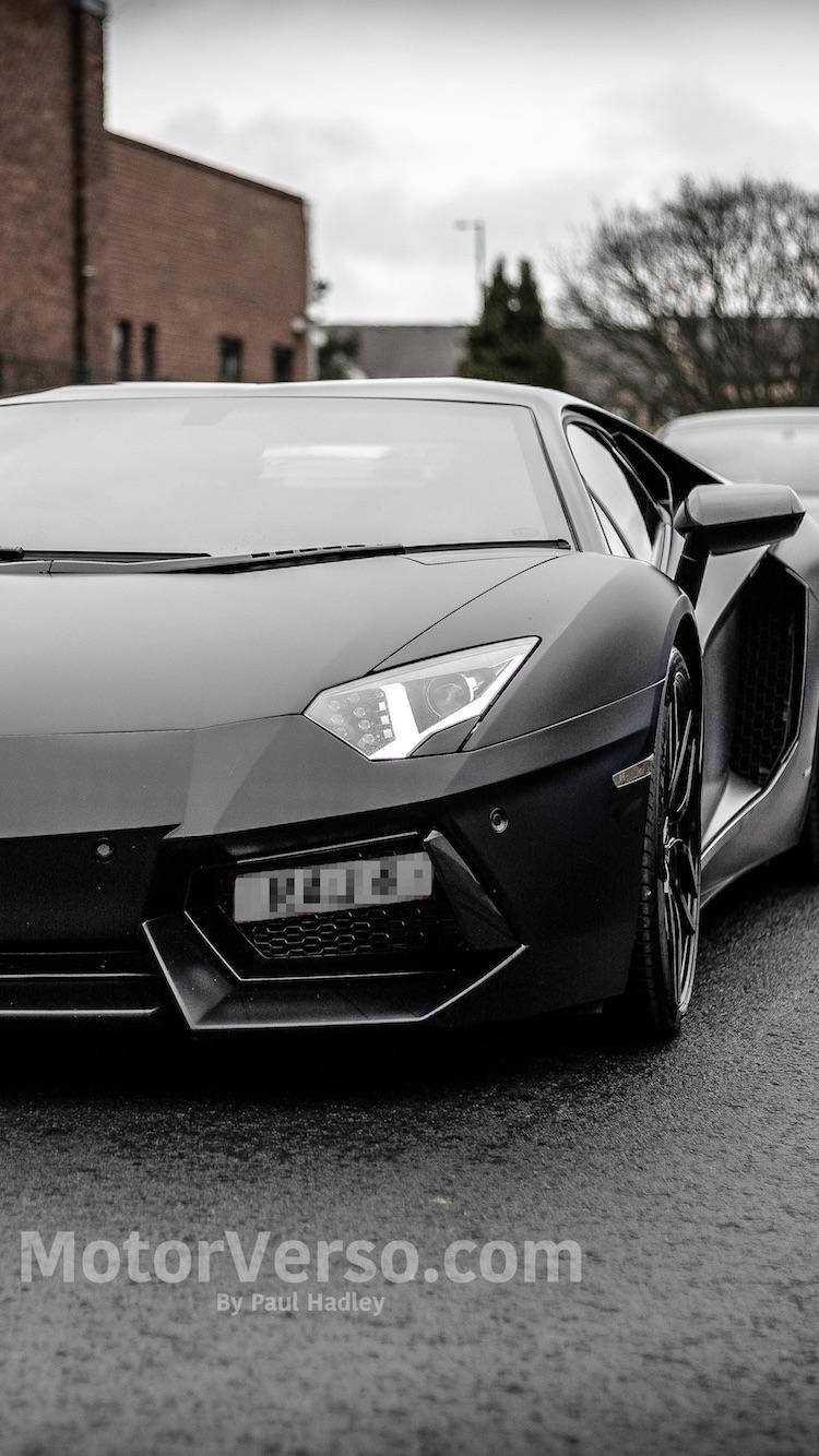 汽車 iPhone 壁紙 桌布 壁紙 — 一台時尚的消光黑色藍寶堅尼Aventador停在濕漉漉的路面上，背景是磚造建築和光禿禿的樹木，天空多雲。