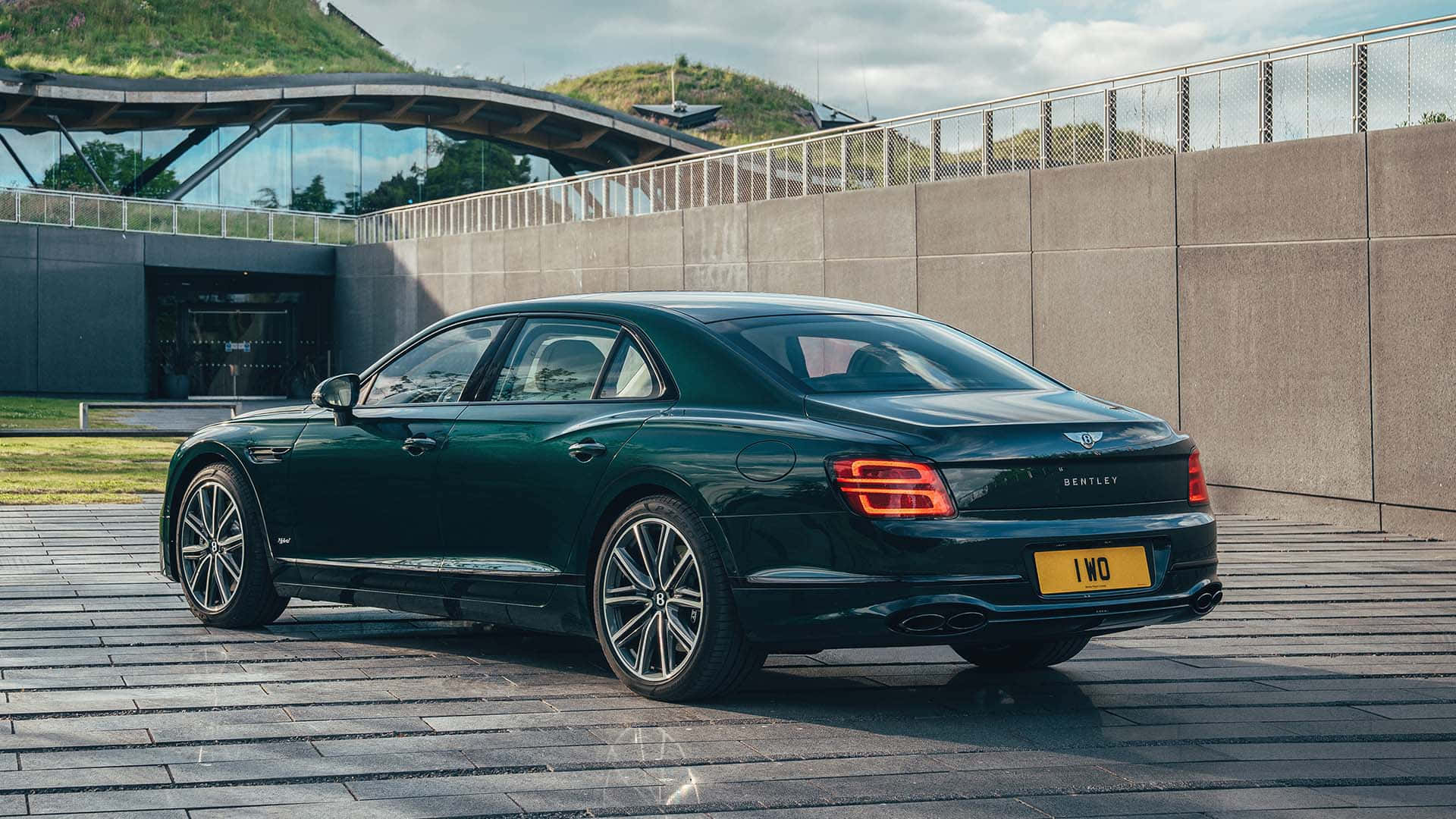 Bentley Flying Spur 桌布 壁紙 — 一台時尚的深綠色賓利飛馳停在石頭廣場上，旁邊是現代玻璃建築，背景有草地山丘。