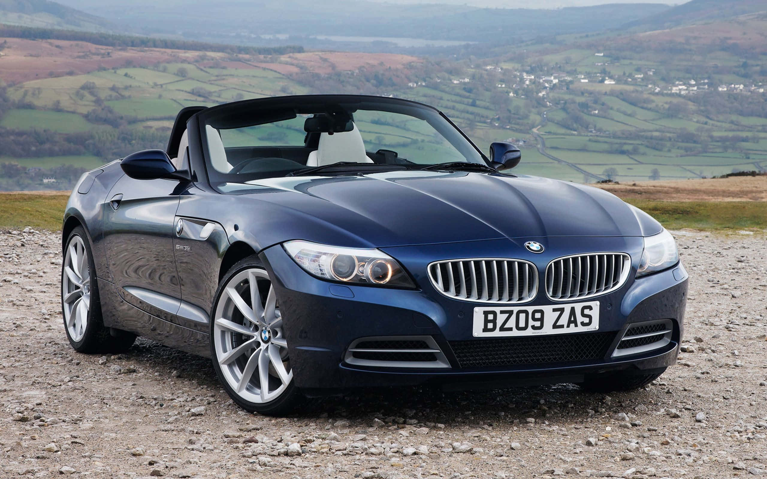 Bmw Cars 桌布 壁紙 — 一台時尚的深藍色BMW Z4敞篷跑車停泊在碎石山頂上，俯瞰著連綿起伏的綠色山丘和遠處的村莊。