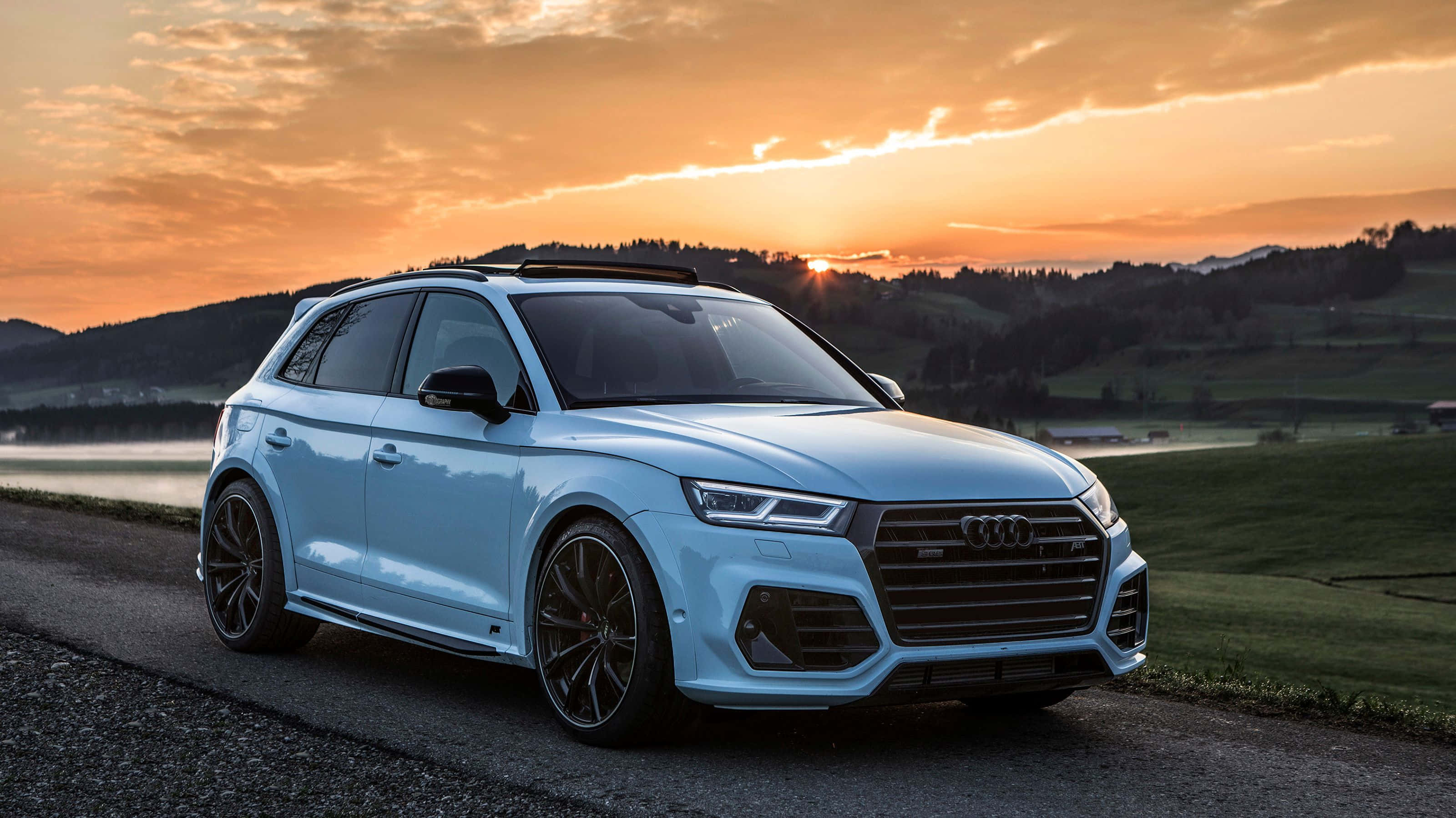 Audi Sq5 桌布 壁紙 — 一台時尚的淺藍色 Audi SQ5 SUV 停泊在日落時分的鄉村道路上，背後是金色的天空和起伏的綠色山丘。